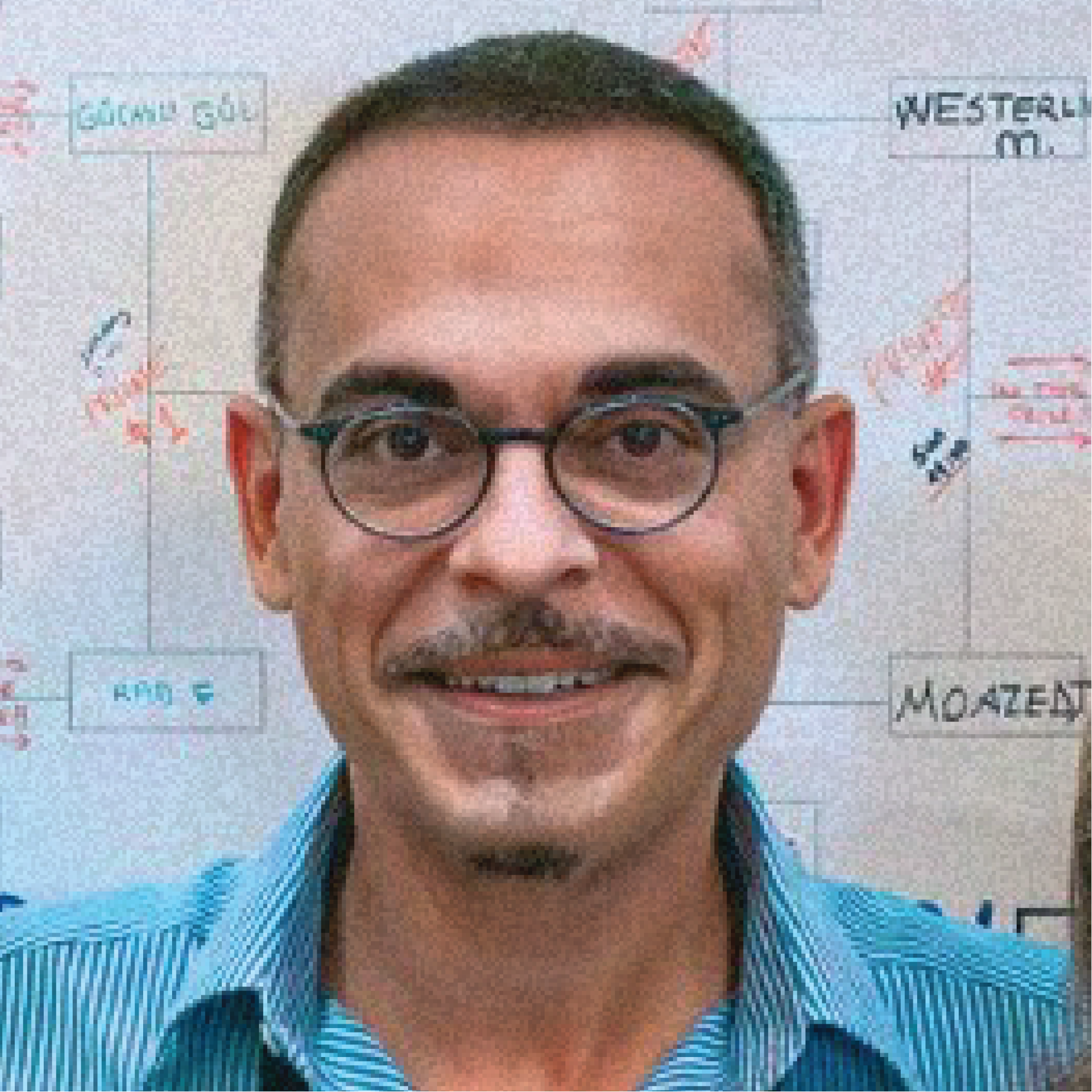 A man with short dark hair, glasses, and a mustache wearing a blue striped shirt, standing in front of a whiteboard with handwritten names and notes.