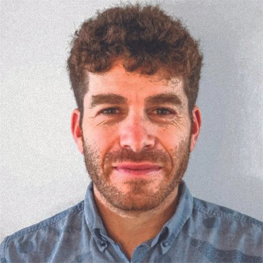 A man with curly brown hair and light skin, wearing a blue button-up shirt, standing against a plain light gray wall.