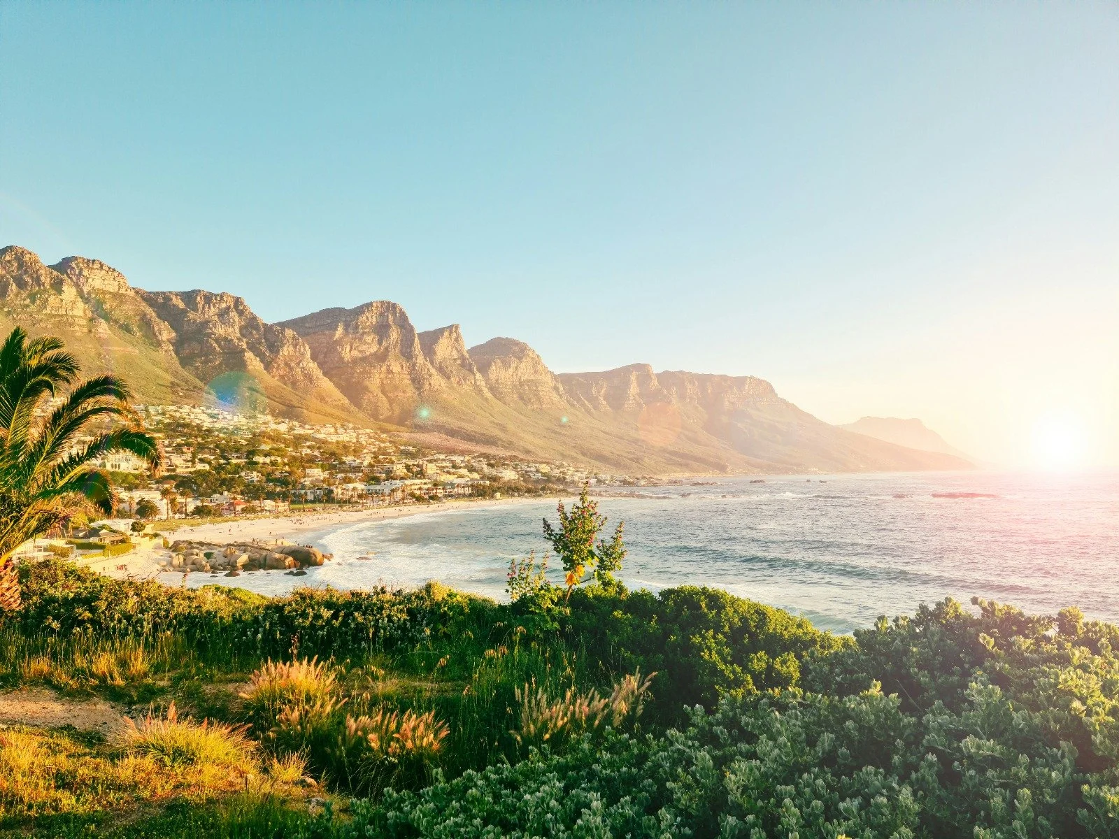 Sunset over a coastal landscape with mountains, a beach, ocean waves, green plants, and a small town in the distance.
