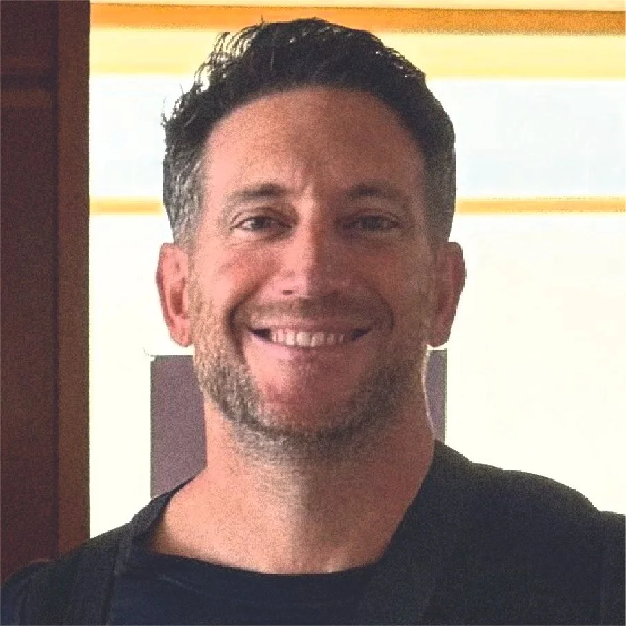 A man with dark, short, styled hair and light facial hair smiling at the camera, wearing a dark shirt with a backpack, standing indoors with light coming from behind.