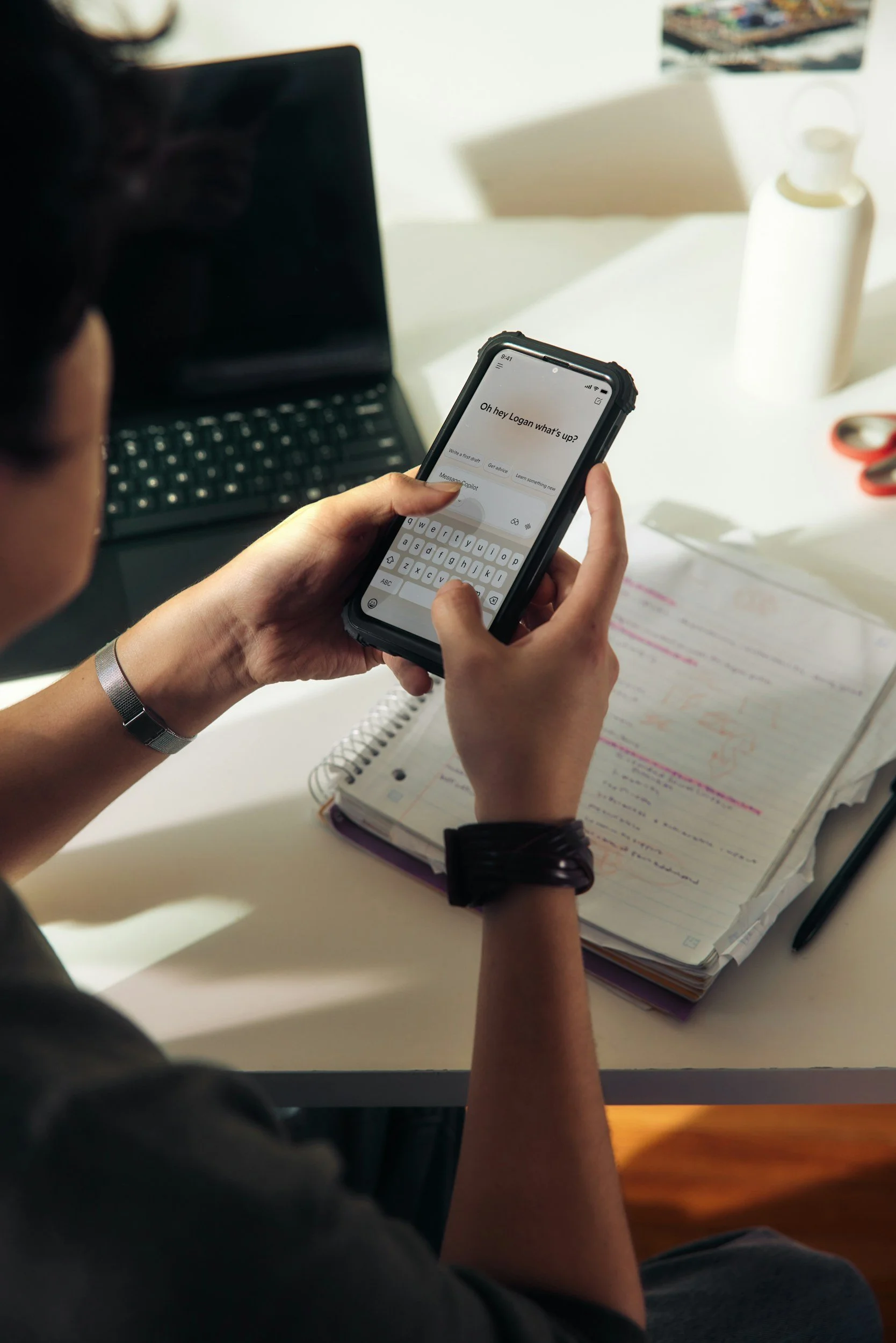 Person holding a smartphone with a messaging app open, windowing a conversation starting with 'Oh hey Logan what's up?'. The person has a spiral notebook with handwritten notes and highlighted text, a laptop, a white bottle, and scissors on a white desk.