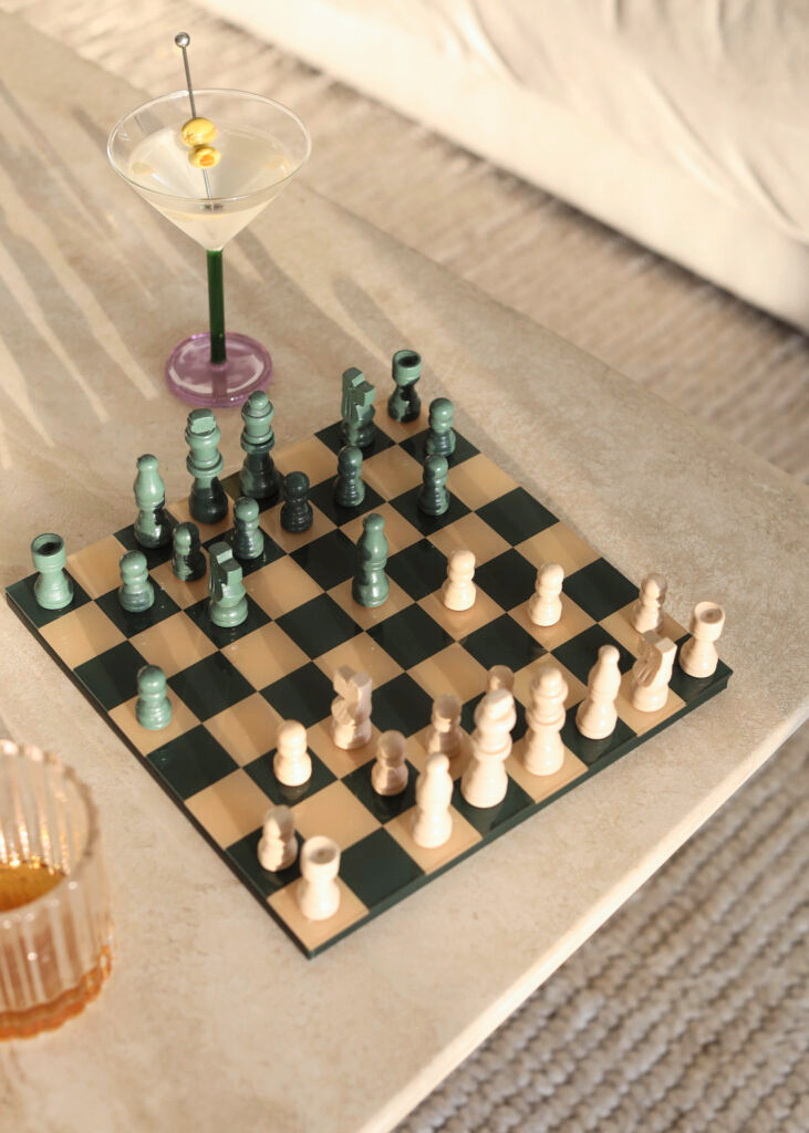 A chess game in progress on a beige checkered board with white and greenish pieces, a green drink with a yellow garnish and cocktail stick on the table, a golden candle holder at the corner, and a beige carpet underneath.
