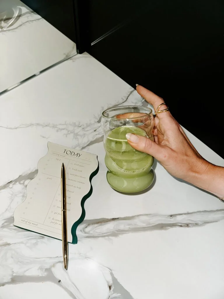 Hand holding a glass with a green beverage on a white marble countertop; planner and gold pen nearby.