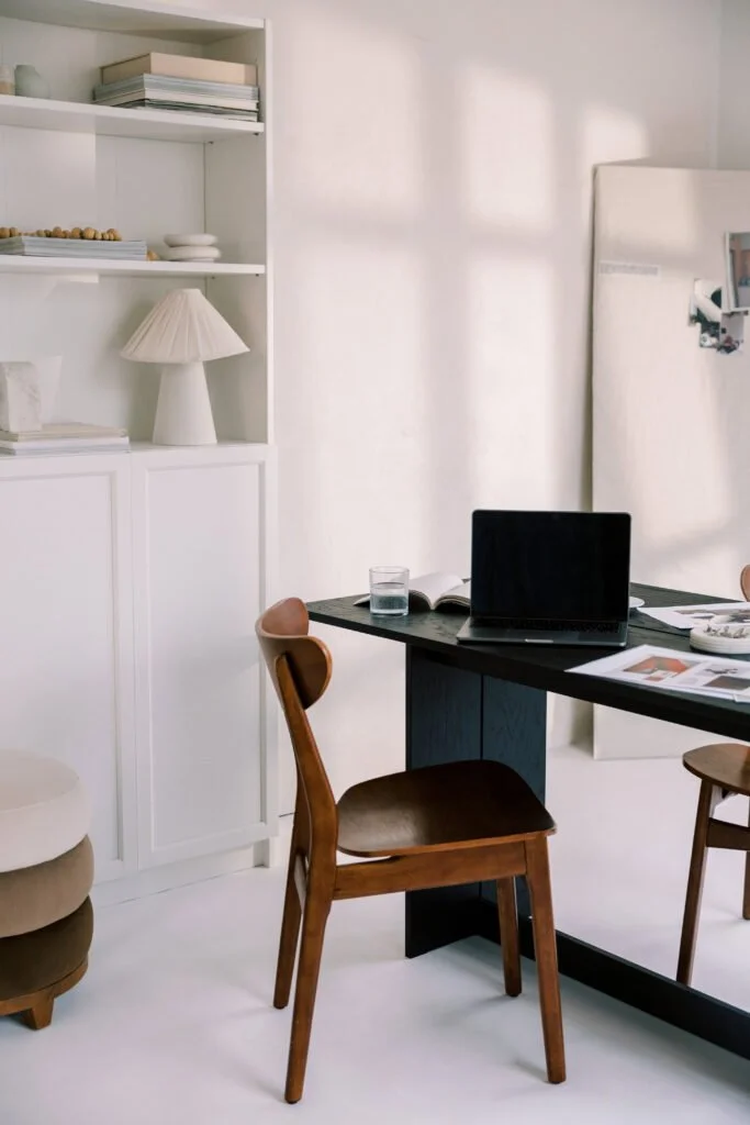 A home office or study area with a dark wooden desk, a wooden chair with a leather seat, a laptop, a glass of water, and some papers. To the left, there is a white shelving unit with books, decorative items, and a lamp. A white board or large mirror is on the right side of the room.
