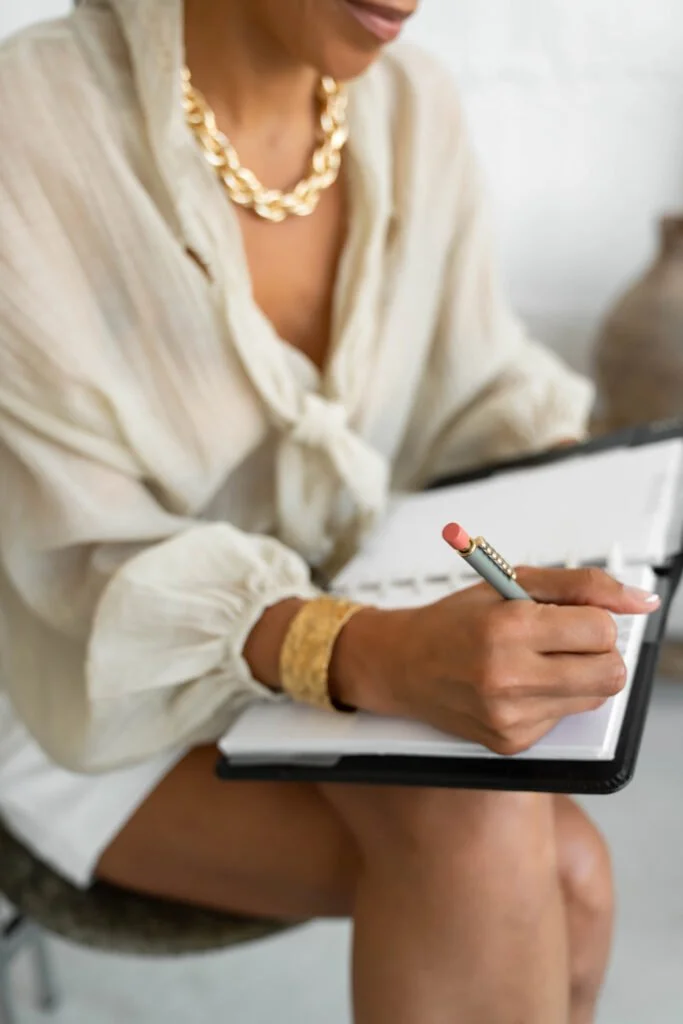 Woman wearing a cream-colored blouse with a gold necklace writing in a notebook with a pink pen.