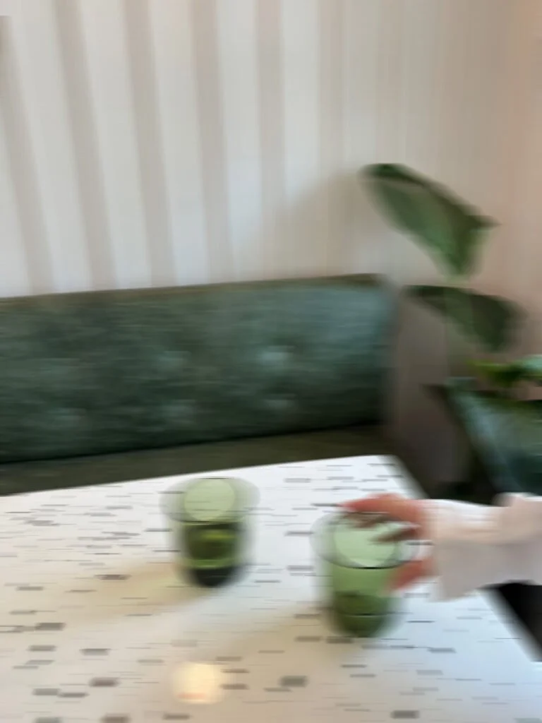 A blurry photo of a table with two glasses of green liquid, a green upholstered bench, and a potted plant with large green leaves in the background.