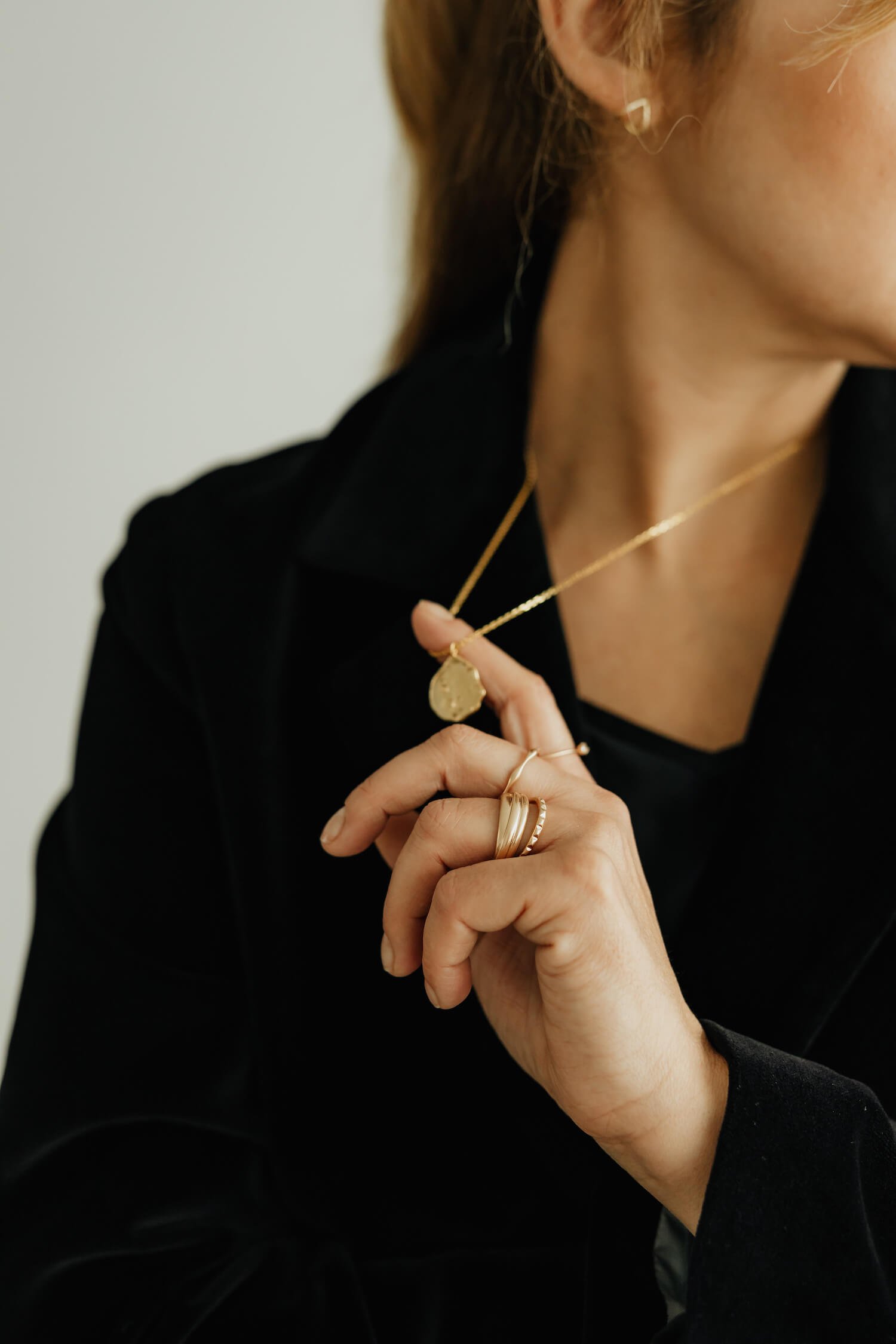 A woman wearing gold jewelry, including a pendant necklace, rings, and earrings, with a black outfit against a plain background.