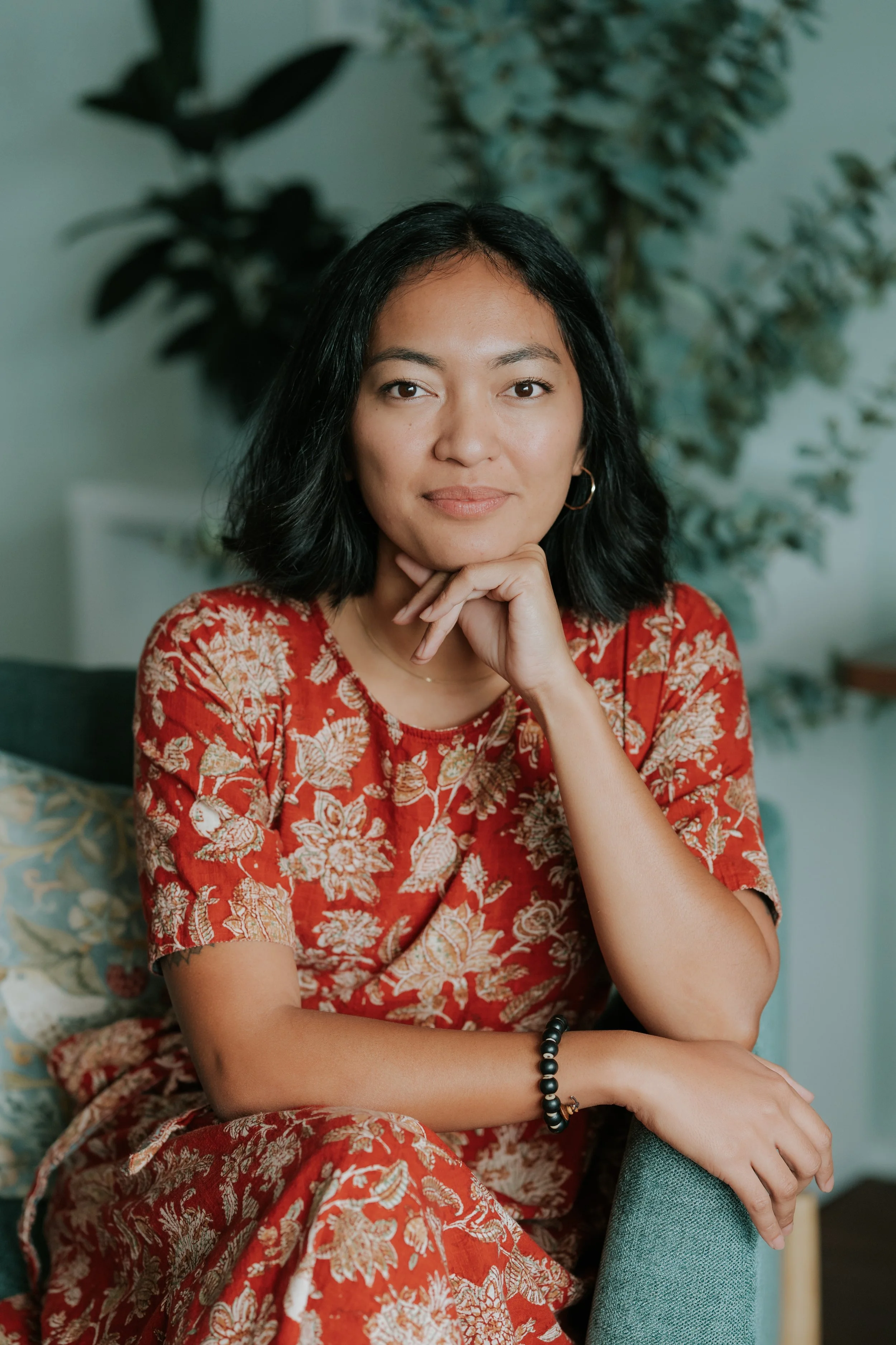 A woman with black hair, wearing a red floral dress, sitting with her hand under her chin, in front of a green background with plants.