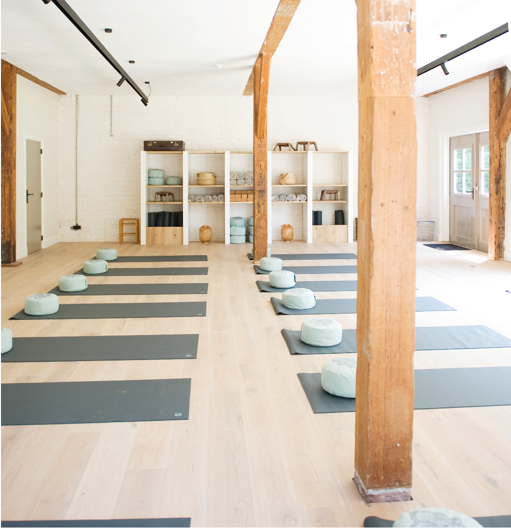 An empty yoga studio with black mats and light green bolsters arranged in two rows, wooden beams, white walls, shelves with towels and storage containers, and a door with glass panels.