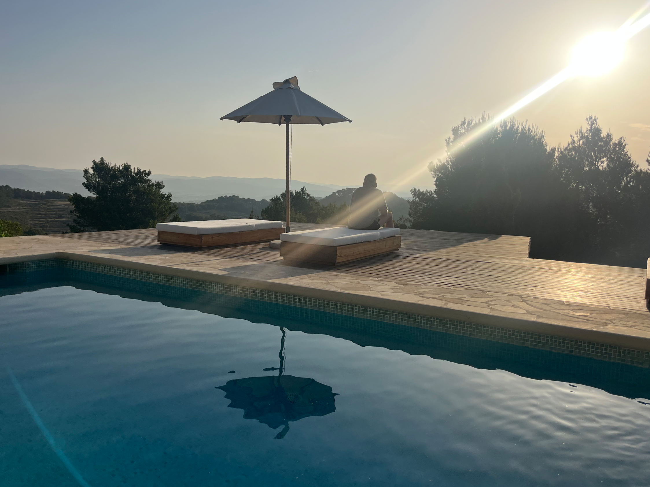 Person sitting on poolside lounge chairs under an umbrella, overlooking a scenic mountainous landscape with trees and a setting sun.