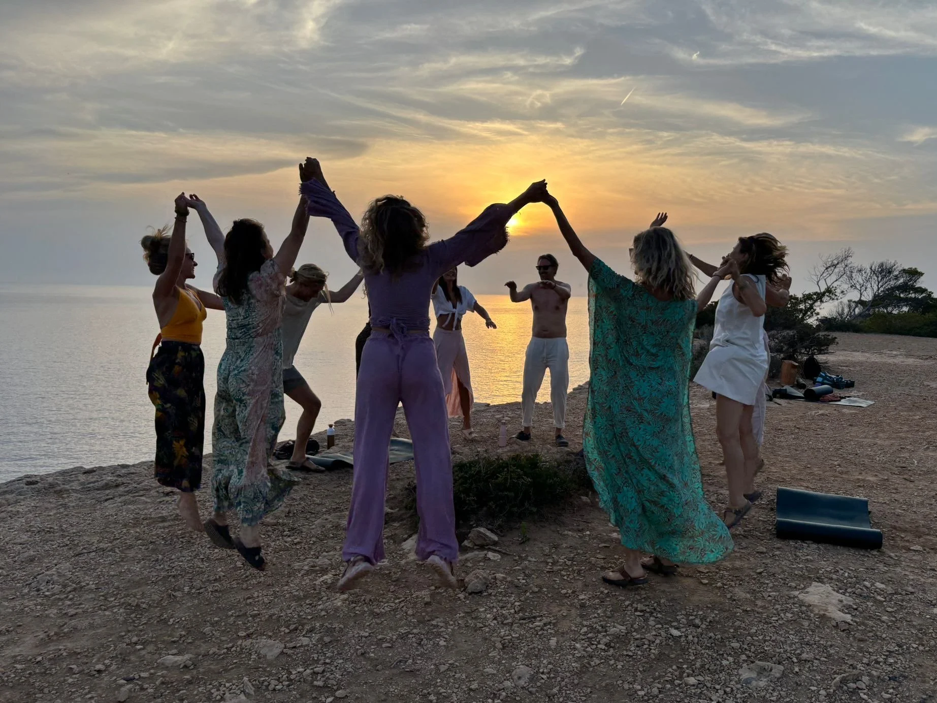 Group of people dancing and celebrating on a rocky shoreline at sunset, with the ocean and sky in the background.