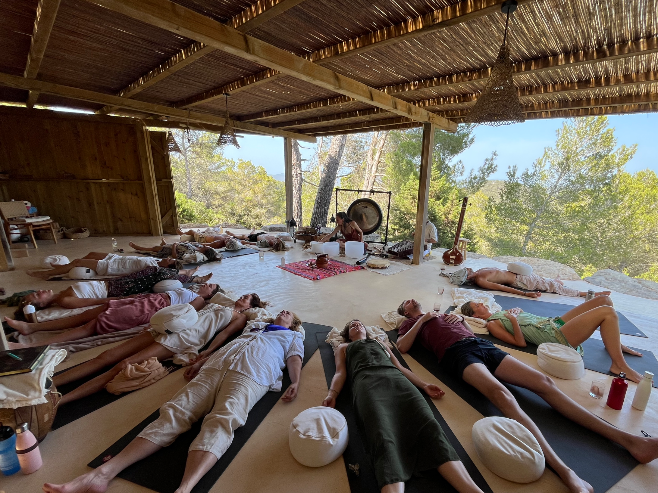 People lying on yoga mats outdoors, participating in a group meditation session under a wooden roof with hanging lamps, surrounded by trees and natural scenery.