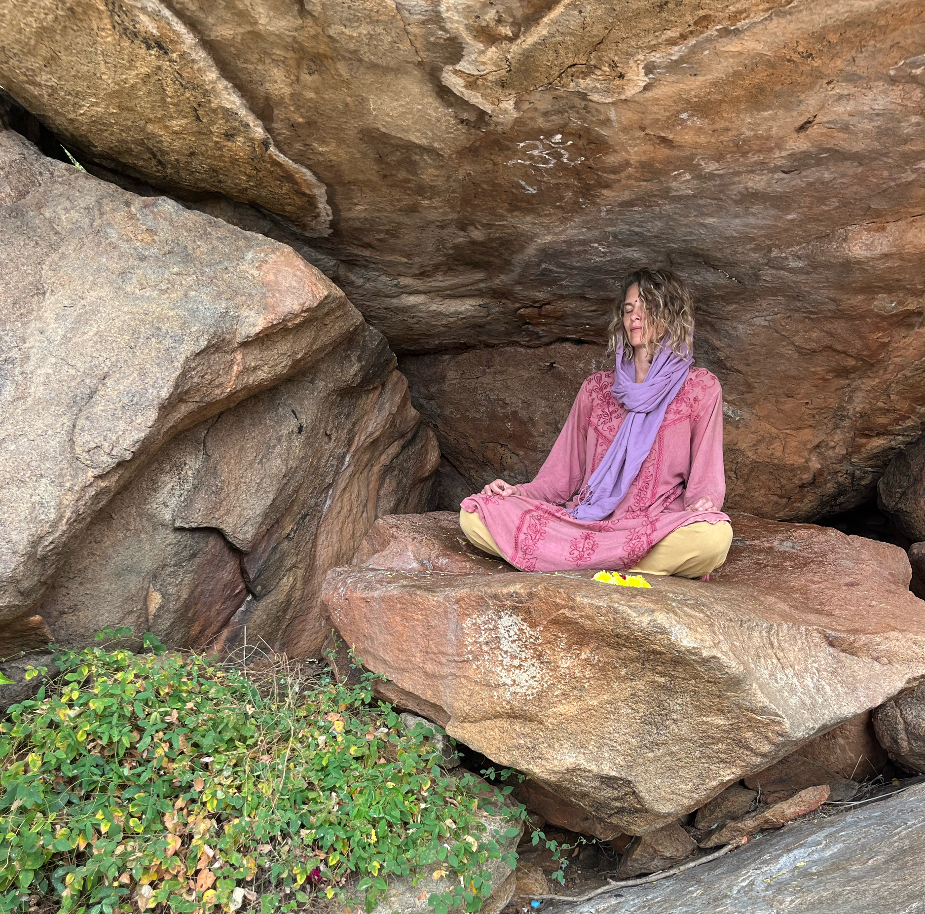 A woman sitting cross-legged in meditation within a small cave, surrounded by large reddish-brown rocks, with some green foliage at the entrance, and white symbols painted on the cave ceiling.