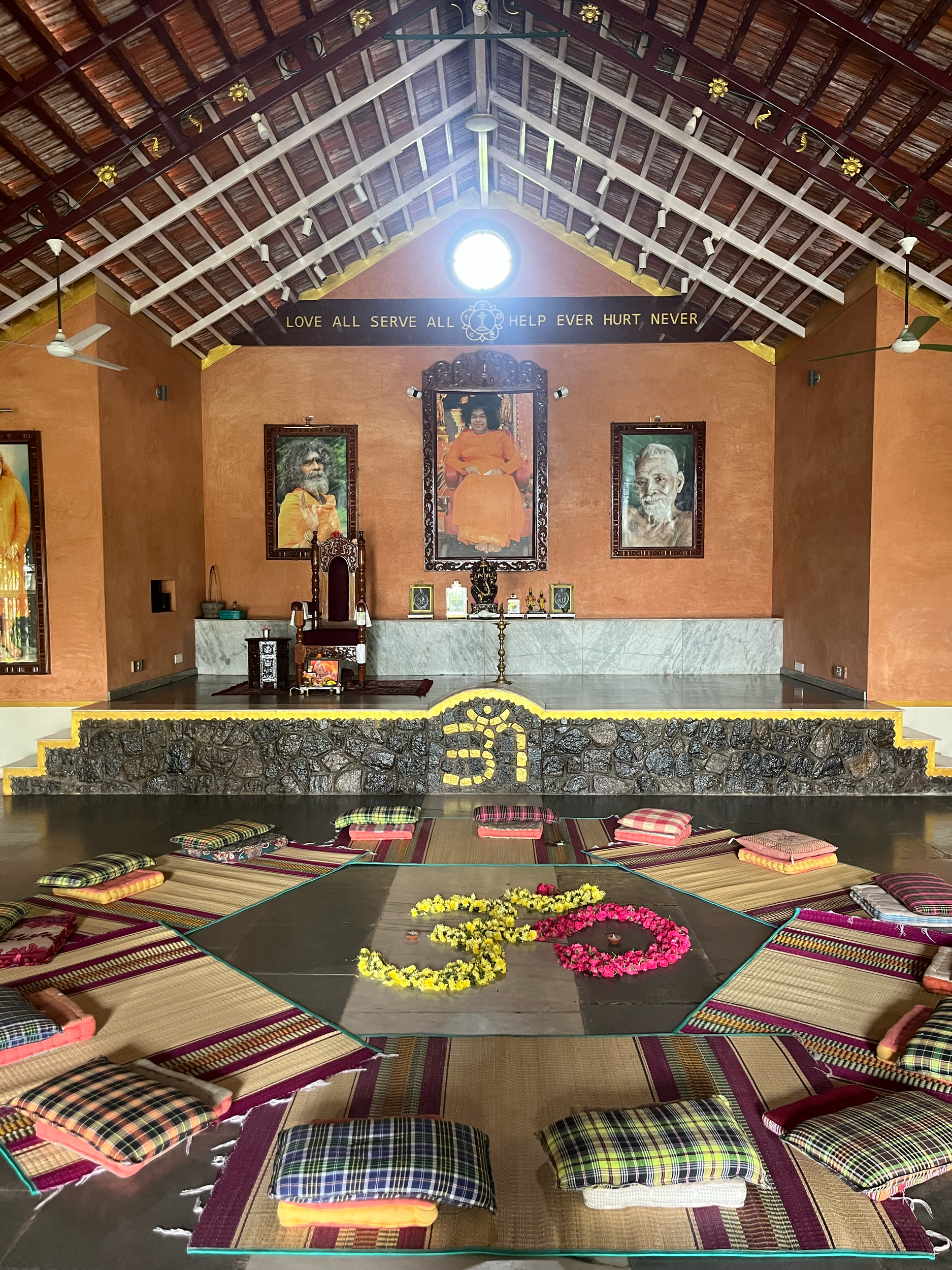 An indoor meditation or prayer hall with cushions on the floor arranged in concentric circles around a central flower decoration. The walls are orange with framed portraits of spiritual leaders and a statue at the center of a raised platform. The cei