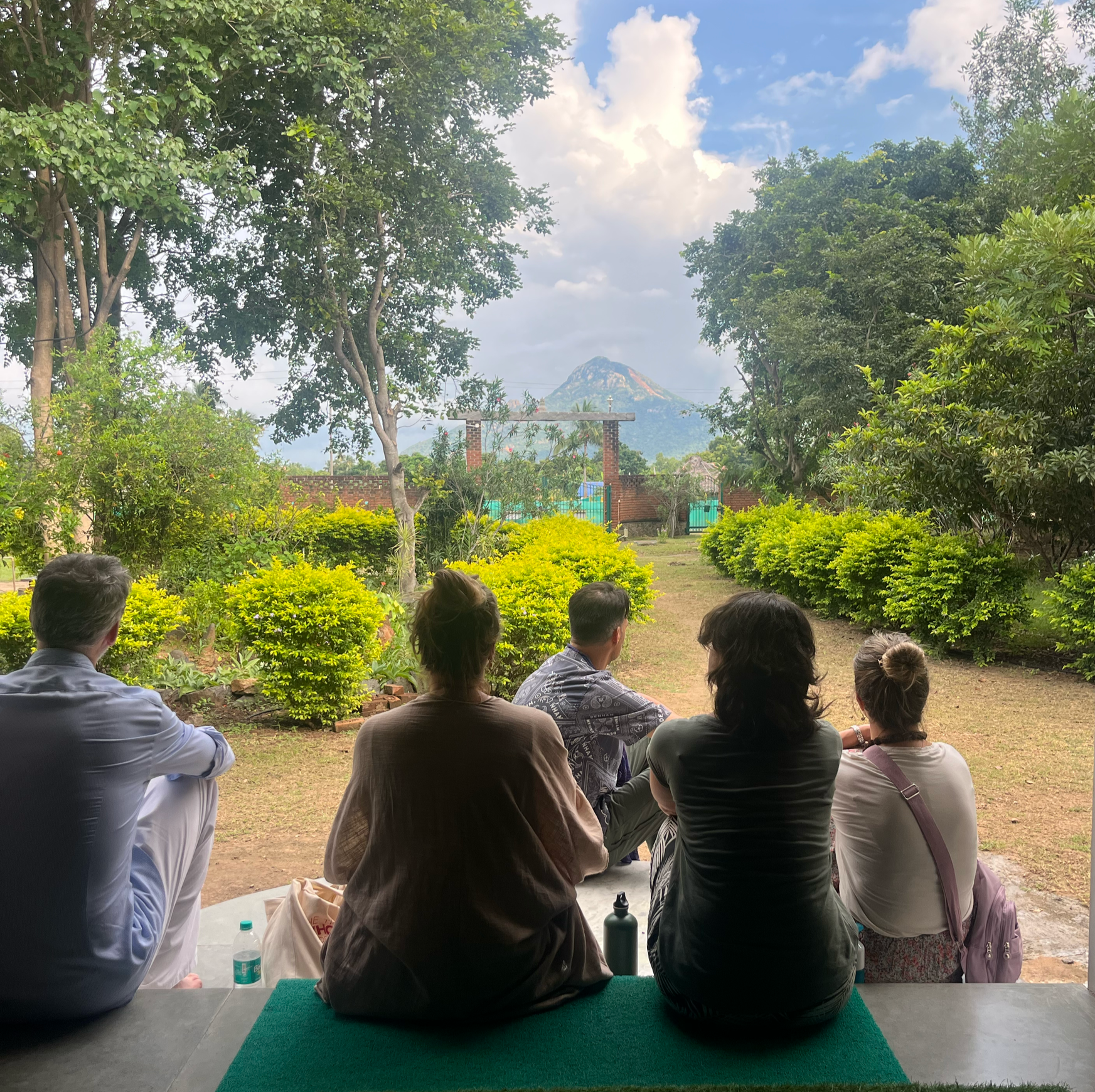 People sitting on benches outdoors, facing a mountain in the distance, with trees and greenery around, and a partly cloudy sky overhead.