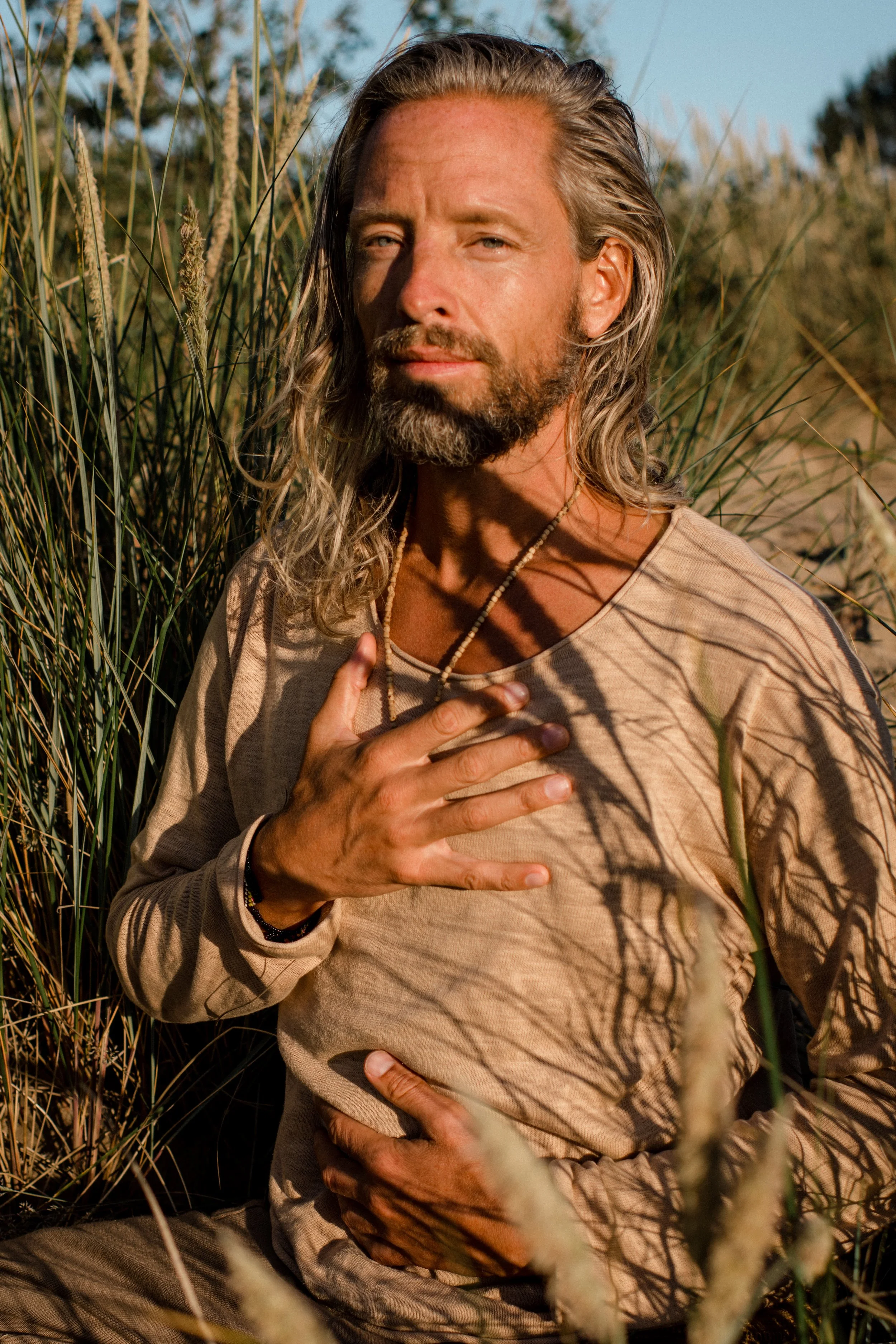 A man with long hair and a beard sitting outdoors among tall grass, holding his chest with one hand and resting the other hand on his abdomen, with shadows cast across his face and body.