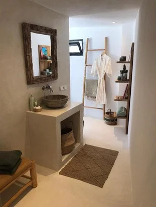 Bathroom with a concrete sink, wooden sink cabinet, wall mirror, small window, wooden shelves with decorative items, and a white robe hanging on a wooden ladder.