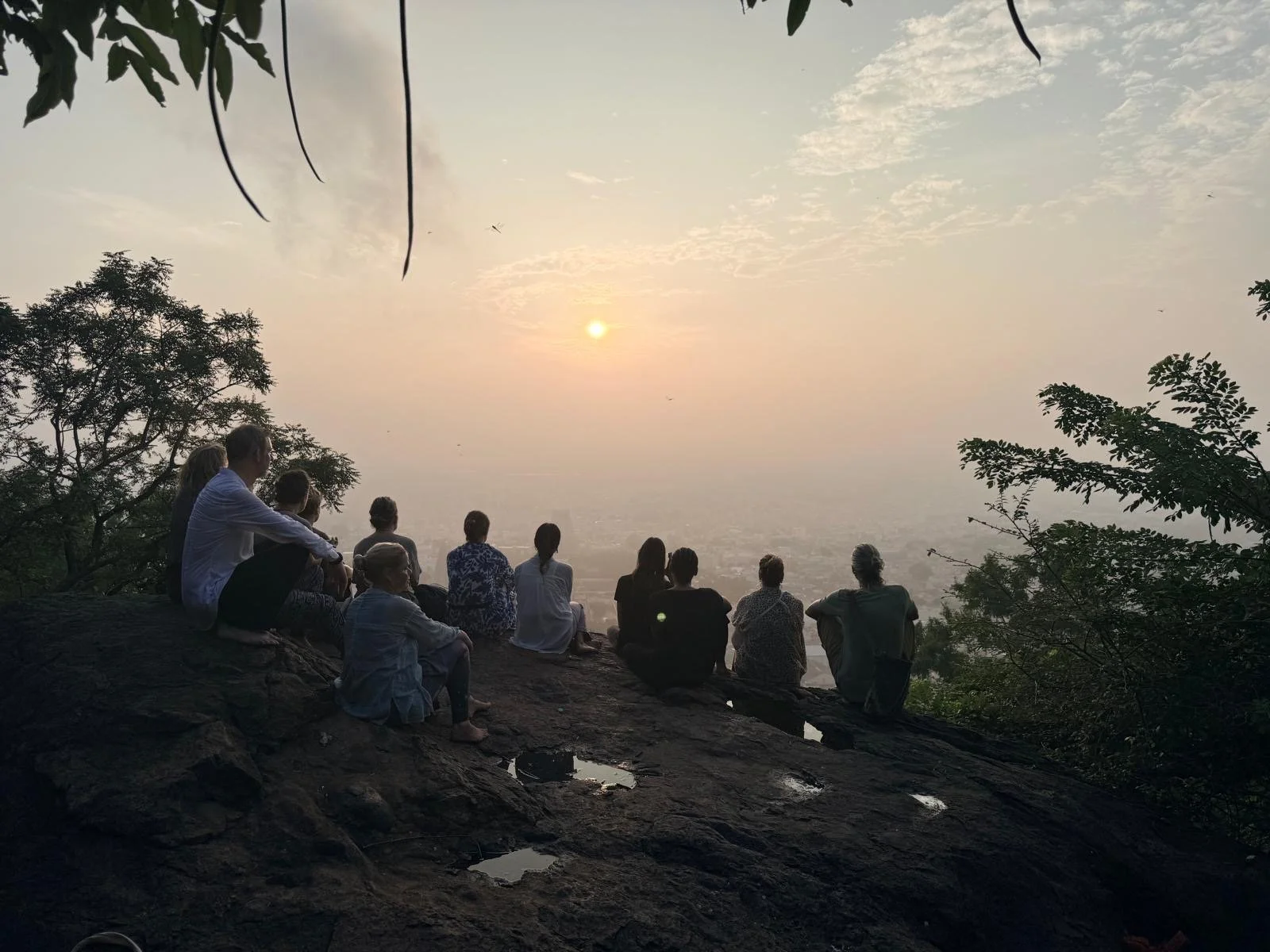 A group of people sitting on rocks and watching a sunset or sunrise from a hilltop with trees around.