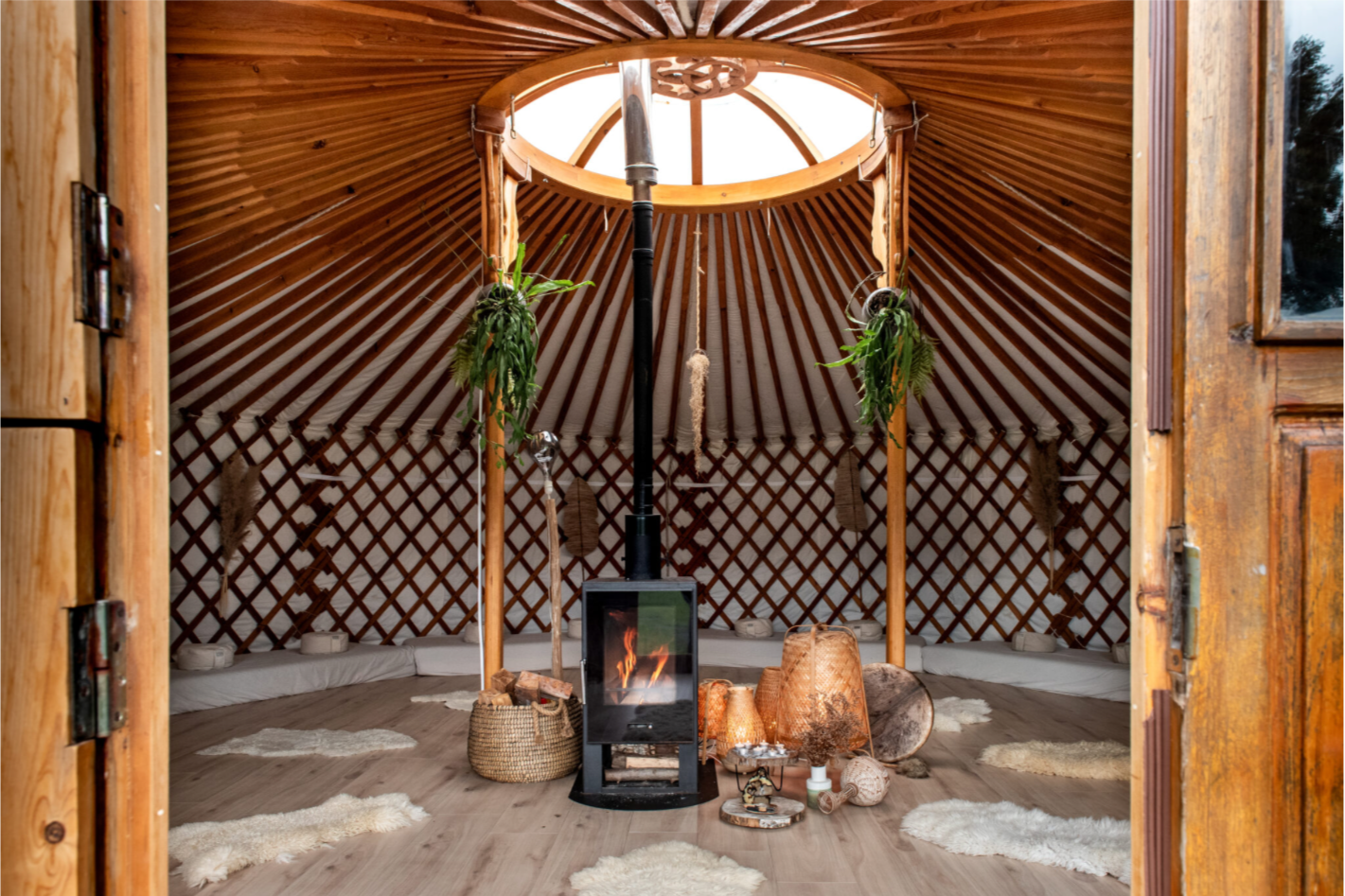 Interior of a cozy yurt with a wood stove, hanging plants, textured lamp, woven baskets, and sheepskin rugs on a wooden floor.