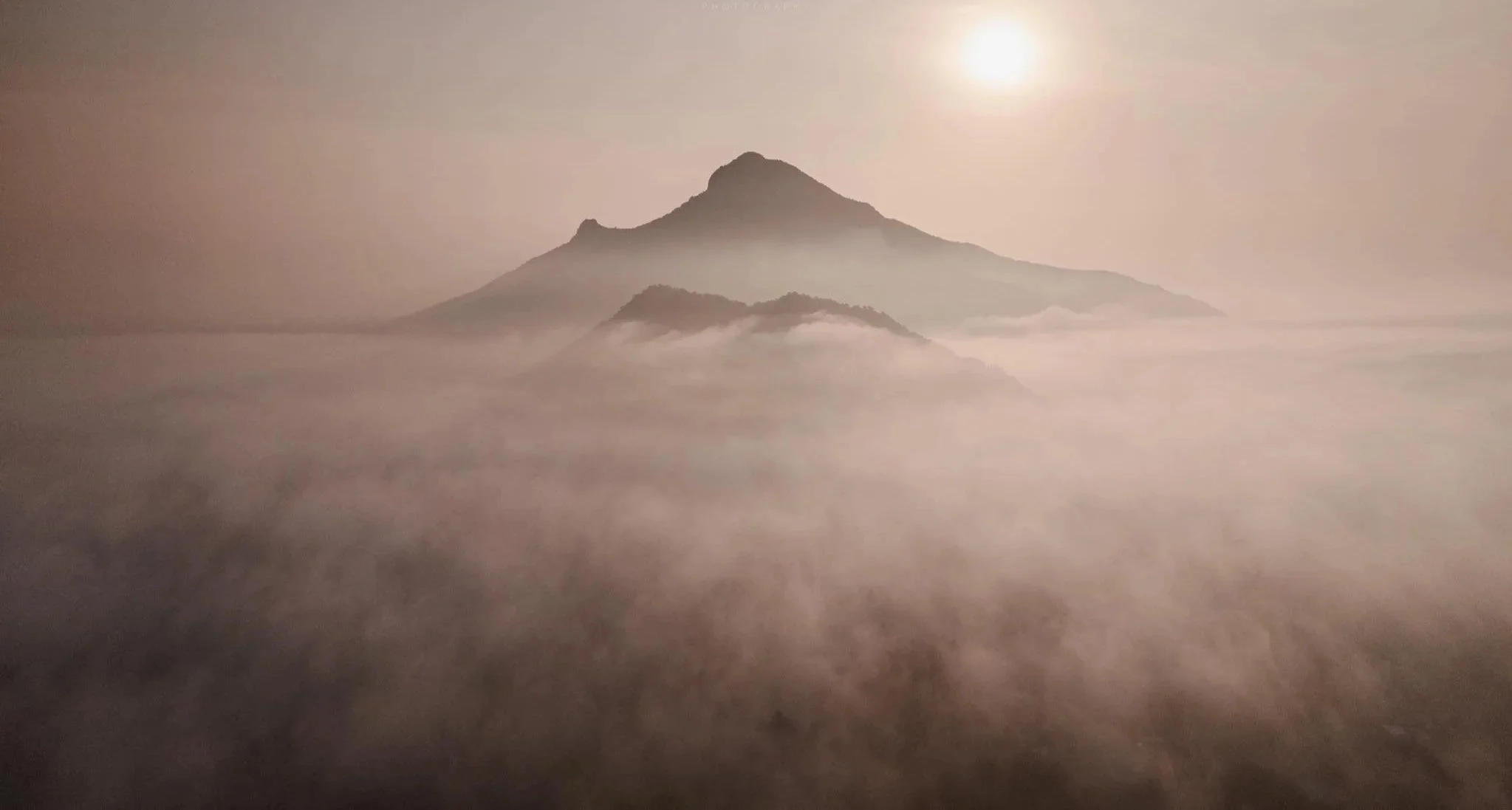 Mountain peak rising above a foggy landscape with the sun shining overhead.
