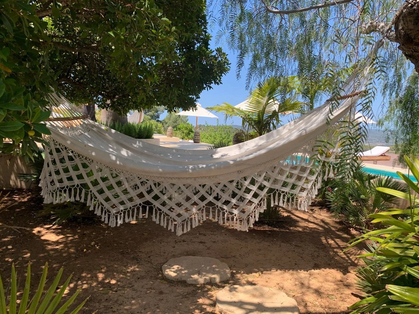 A white hammock with fringes hanging between trees with lush green leaves, near a swimming pool and sun loungers under umbrellas against a blue sky.