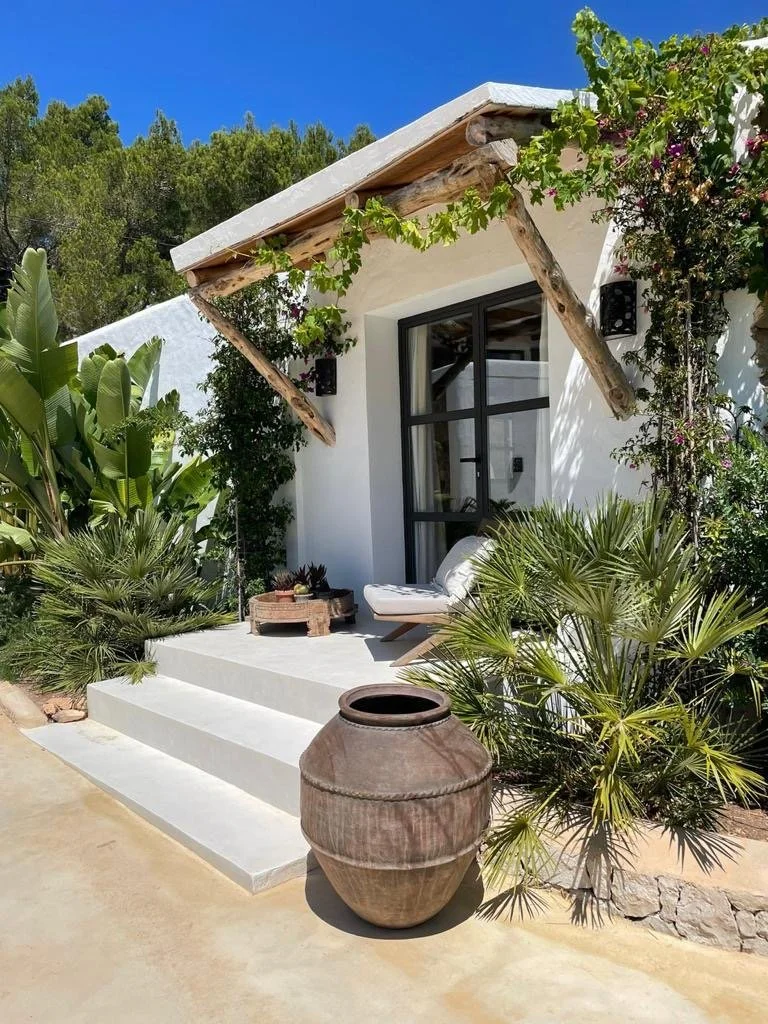 A cozy outdoor patio area with white steps, a lounge chair, and decorative plants in front of a white house with a black-framed door. An large ceramic pot is on the ground near the steps, and a rustic wooden table is on the patio, under a sloped roof with exposed wooden beams.