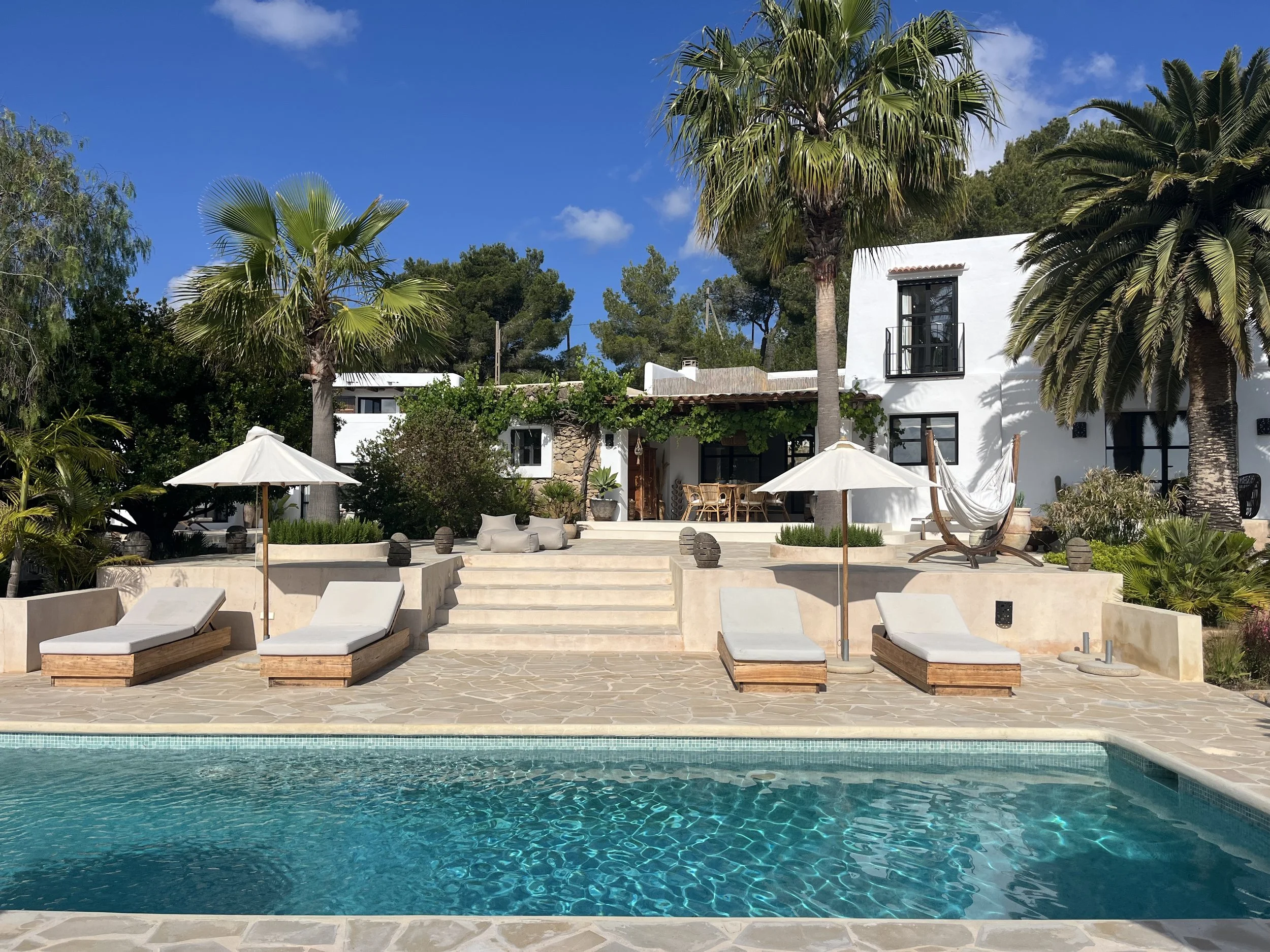 Swimming pool with clear blue water in the foreground, surrounded by stone patio and lounge chairs with umbrellas; white house with black-framed windows and palm trees in the background under a partly cloudy sky.