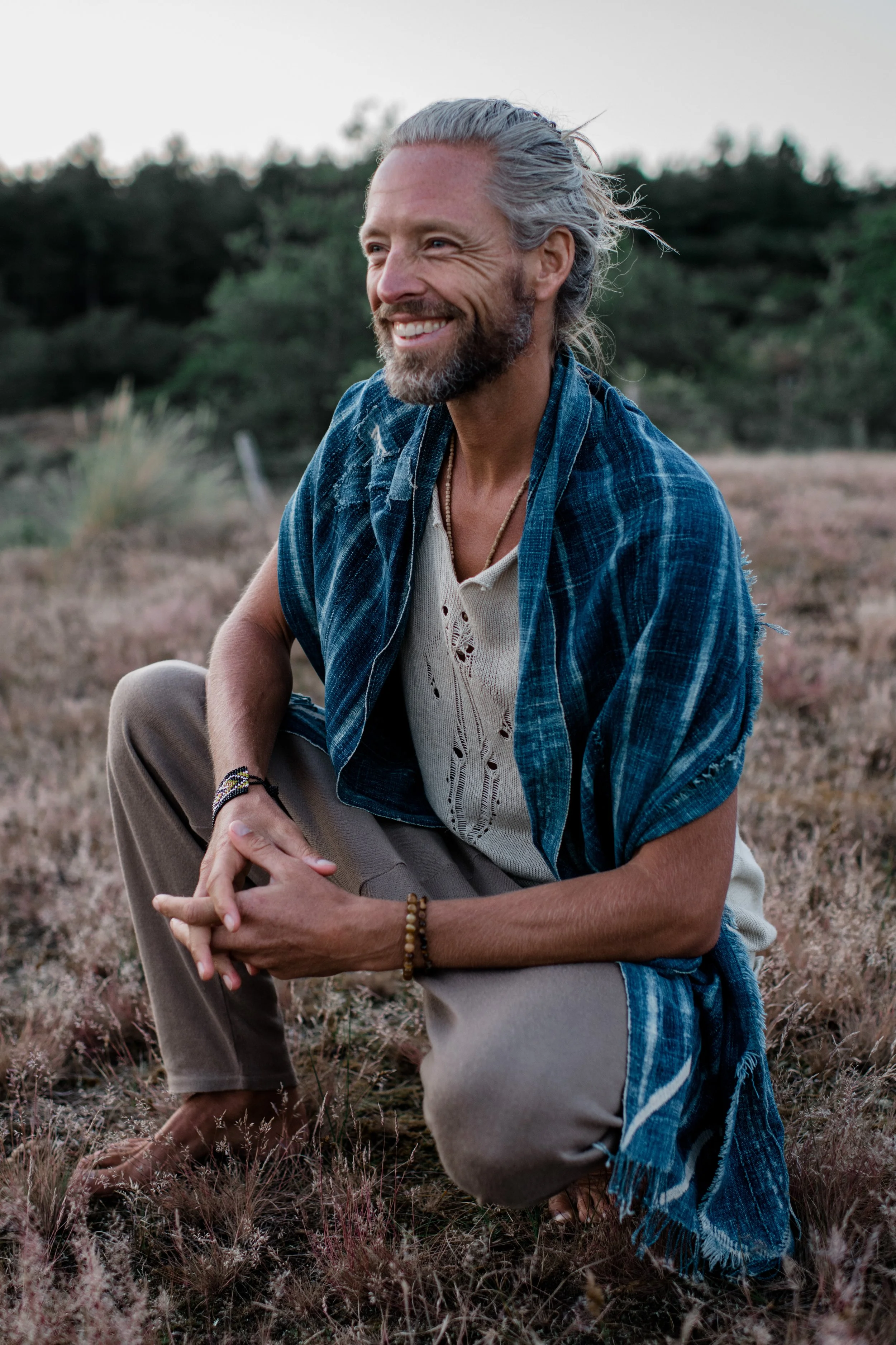 A man with gray hair and a beard smiling in a field with purple grasses, wearing a blue plaid shirt and tan pants.