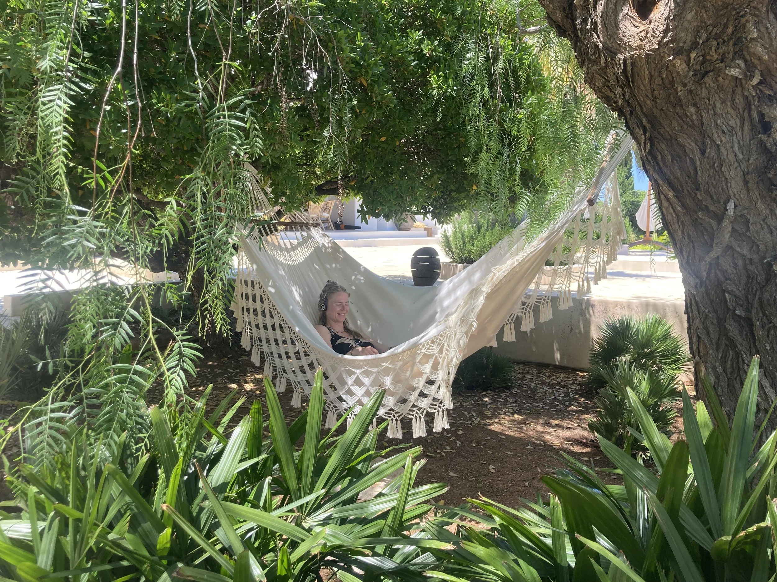A woman relaxing on a hammock hanging between trees in a lush garden with various green plants and a patio area visible in the background.