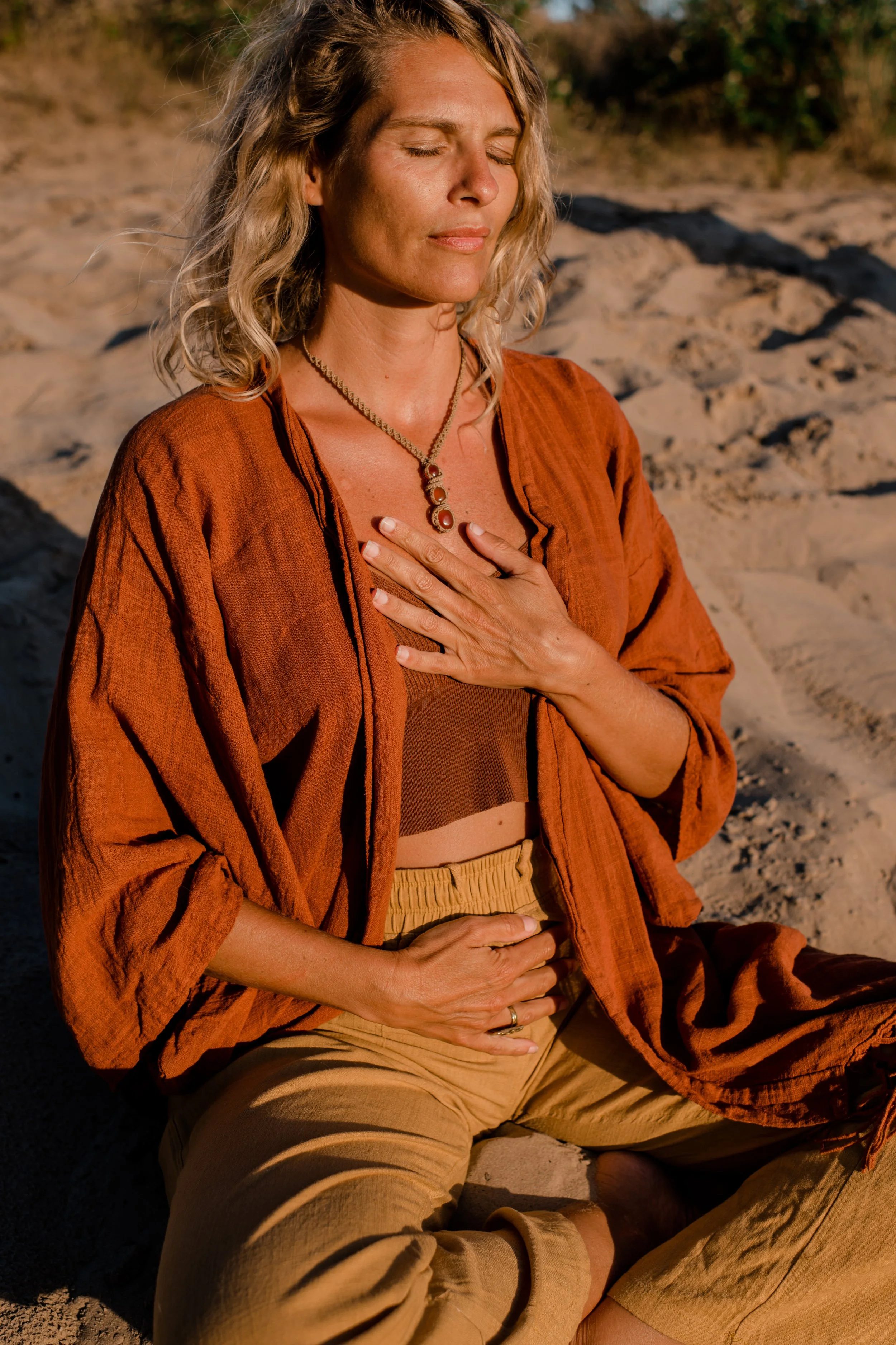 A woman sitting on the sand outdoors with eyes closed, wearing an orange shirt and beige pants, with one hand on her chest and the other on her stomach, in a peaceful moment during sunset.