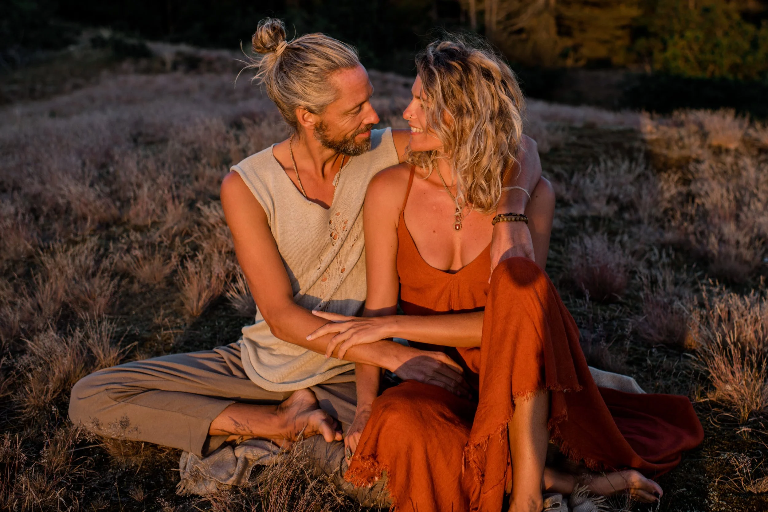 A couple sits closely on the ground outdoors at sunset, gazing into each other's eyes and smiling. The man has long gray hair tied up in a bun and a beard, wearing a sleeveless top and patterned pants. The woman has curly blonde hair, multiple necklaces, and wears an orange dress. They are surrounded by natural grass and rocks, with a blurred background of trees.