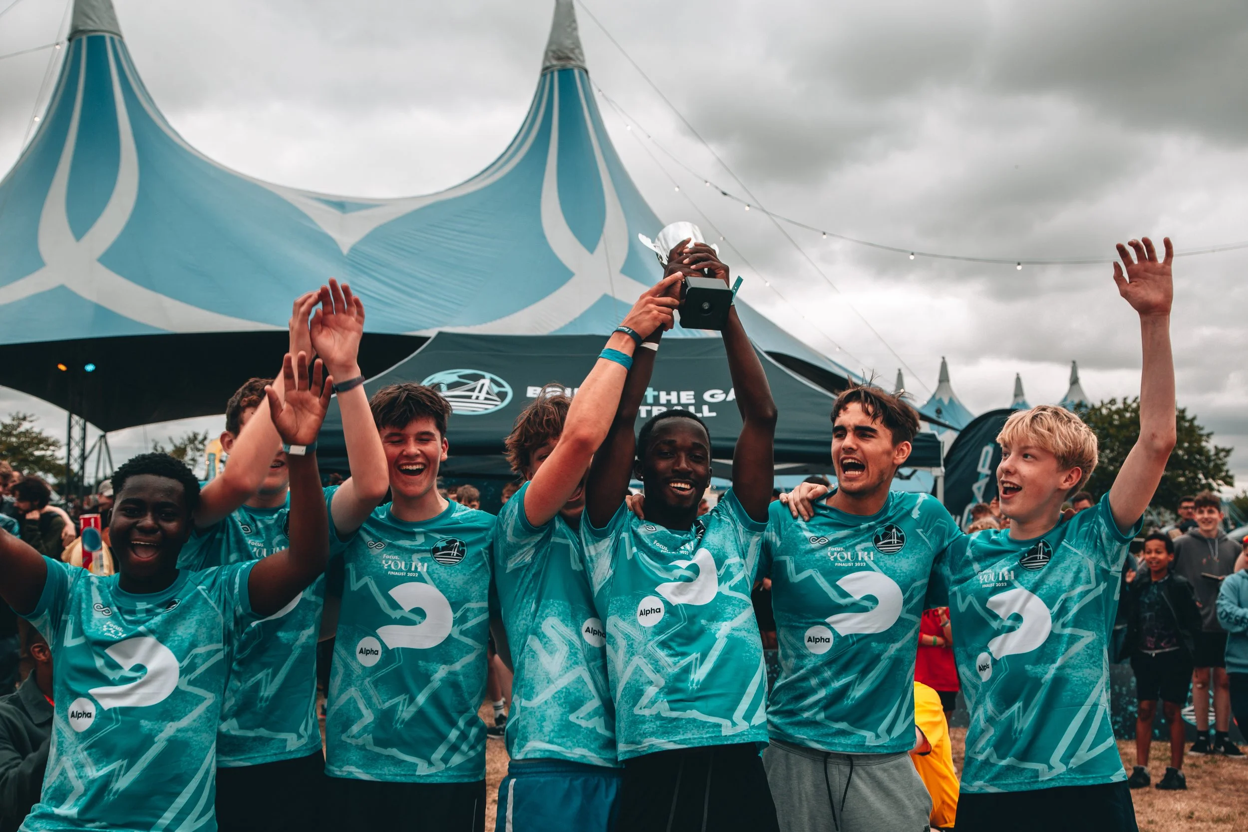 A group of young male soccer players celebrating with a trophy at an outdoor event on a cloudy day.