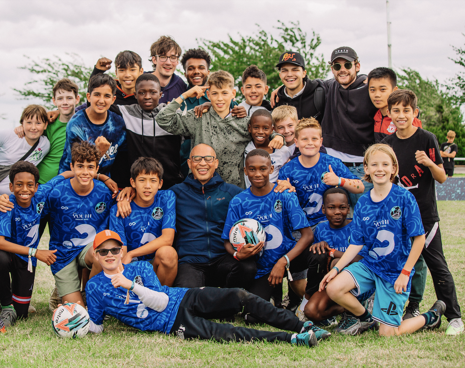 Group of young boys and a few adults, some wearing blue sports jerseys, posing outdoors on a grassy field after a soccer game.