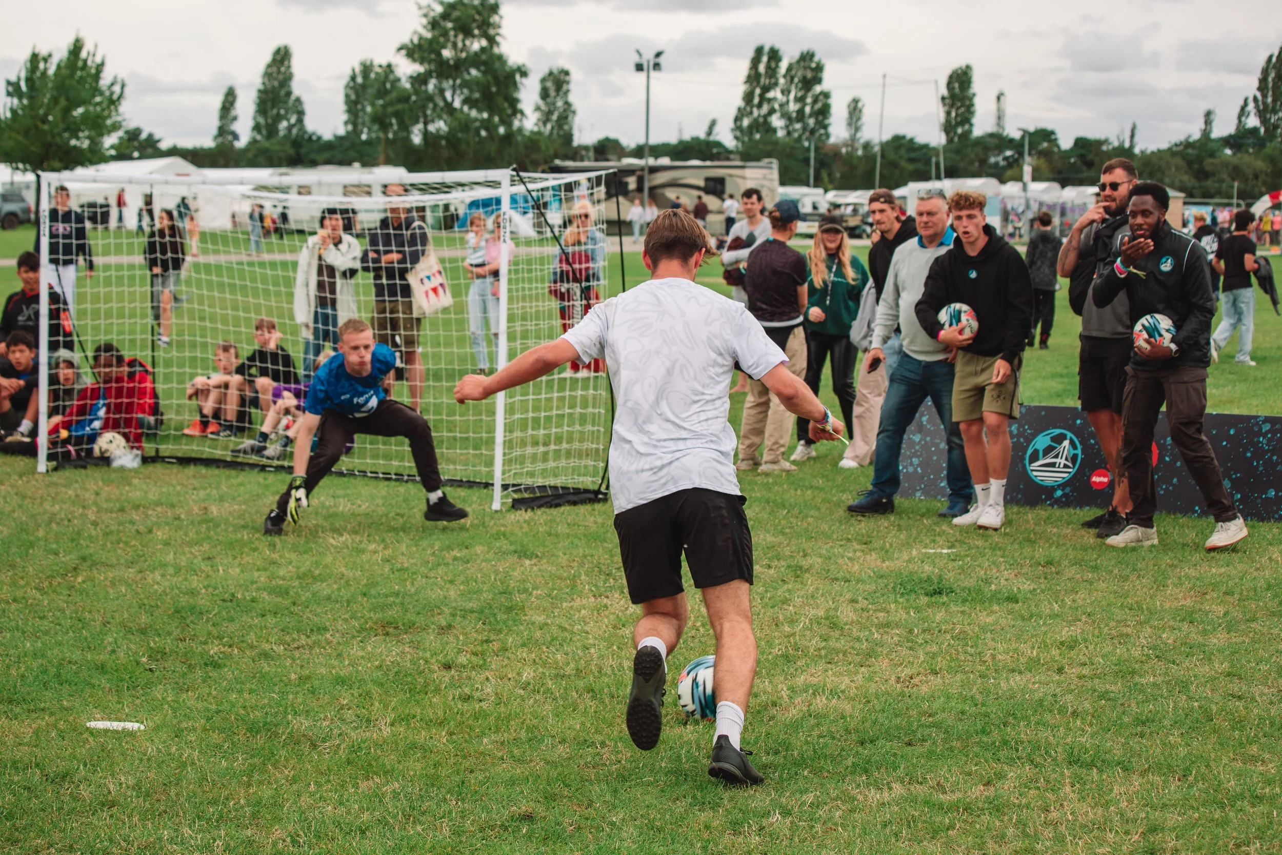 A person in a white shirt and black shorts kicking a soccer ball towards a goal, with a goalkeeper in a blue shirt ready to block. Several spectators and photographers are watching, some holding soccer balls. The scene is set on a grassy field with tents and trees in the background.