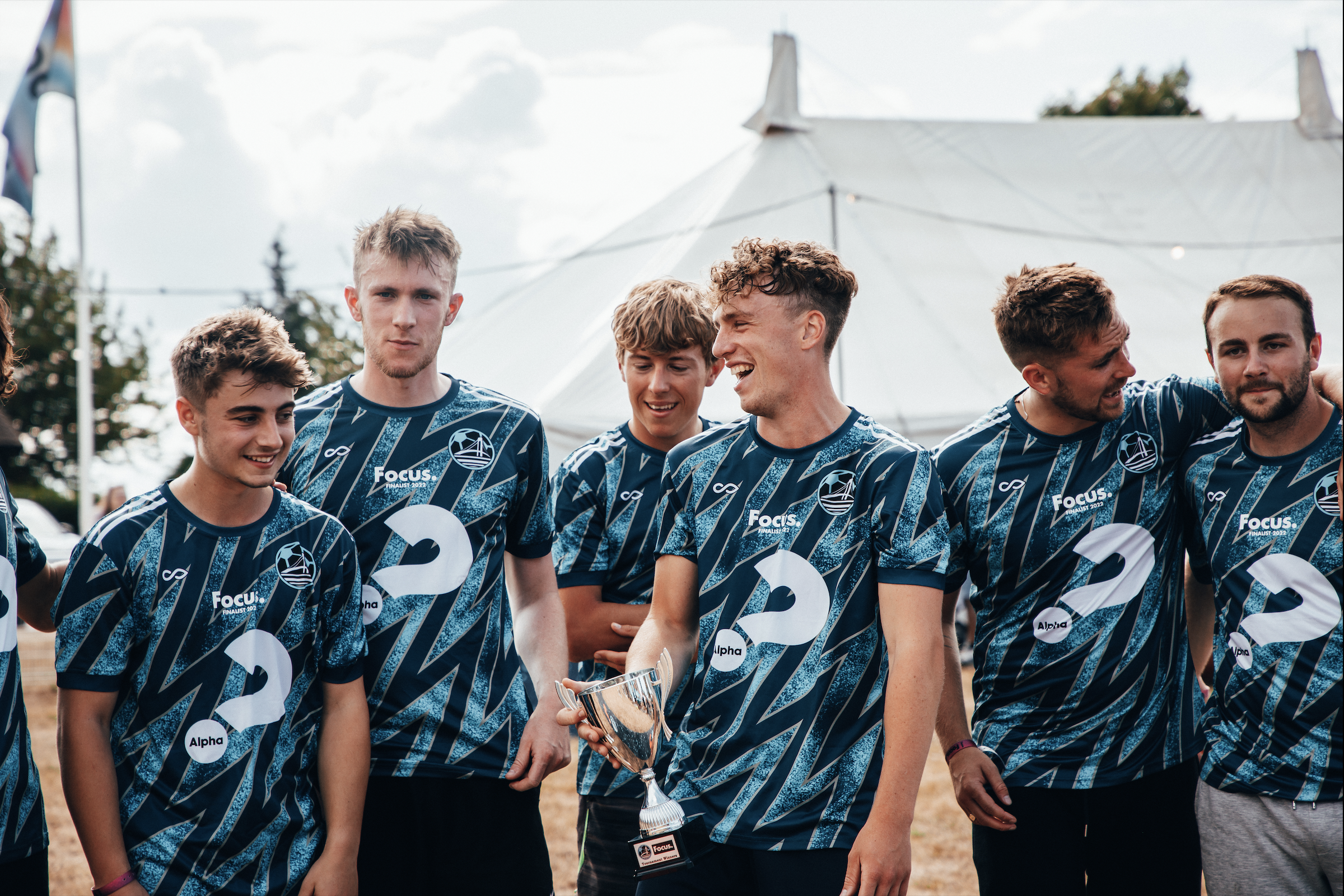 Group of young men in matching sports jerseys standing outdoors, some smiling and one holding a silver trophy cup, with a tent and trees in the background.