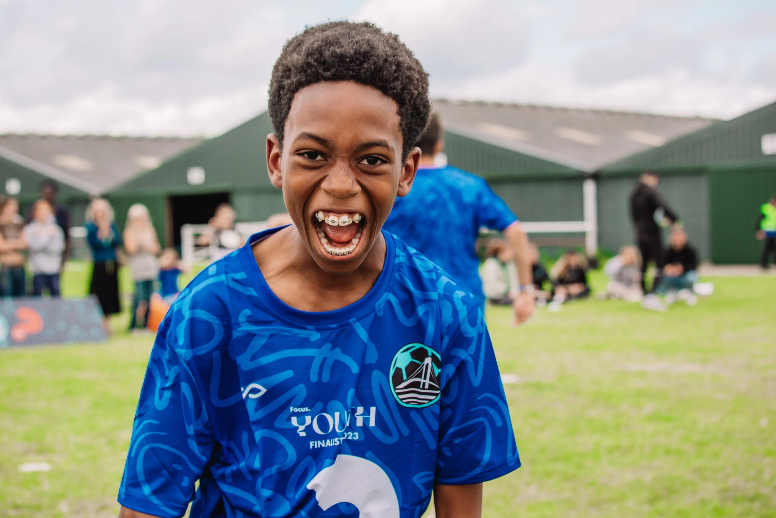 Young boy with braces smiling on a sports field, wearing a blue sports jersey, with other children and adults in the background.