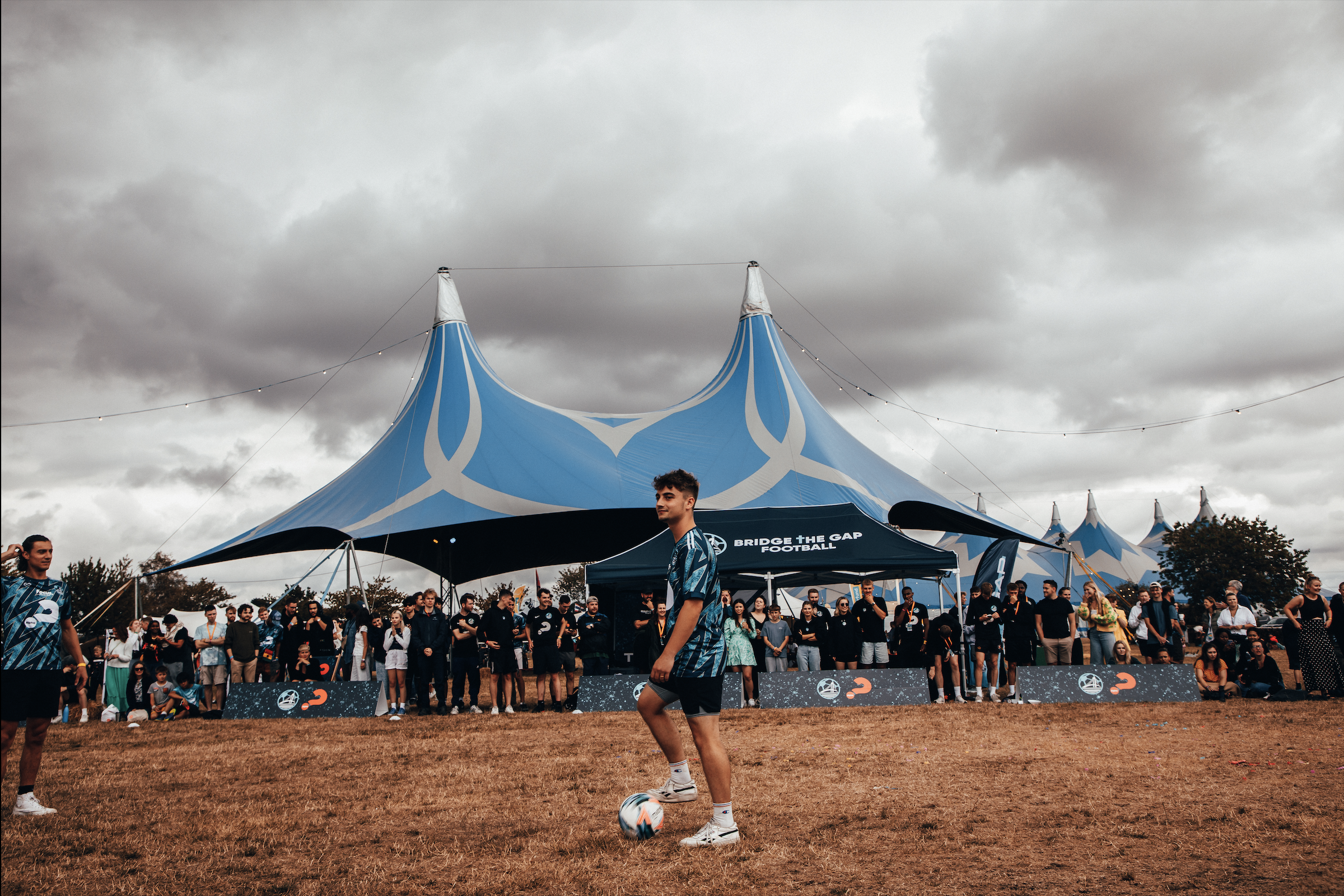 A young man standing on a soccer field with a ball in front of a crowd under a large, blue, tent-like structure at a sports event.
