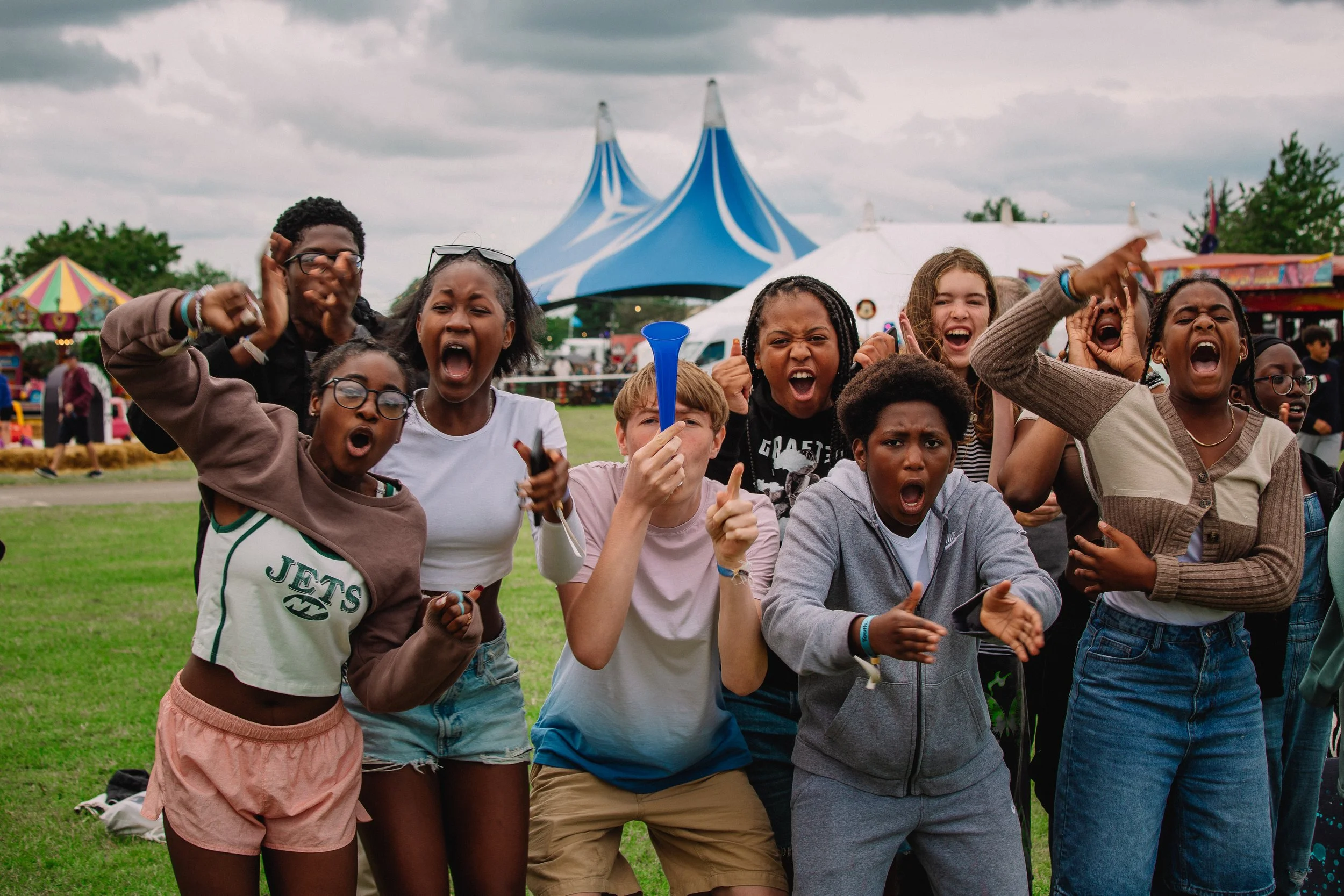 A group of children at an outdoor festival, appearing excited and shouting, with tents and carnival rides in the background.