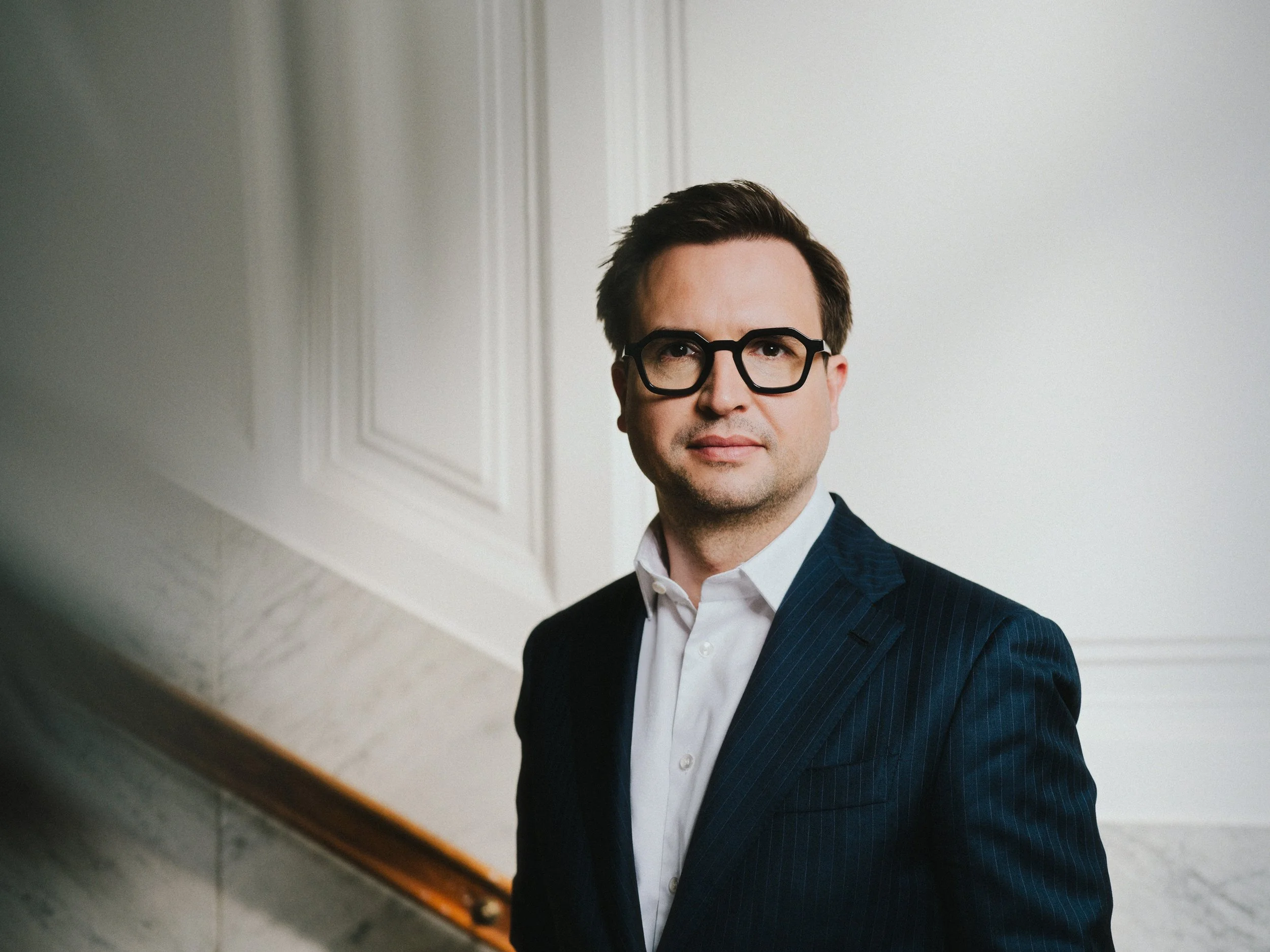 Ein Mann mit dunklen Haaren, Brille und Anzug steht vor einer weißen Wand in einem eleganten Raum.