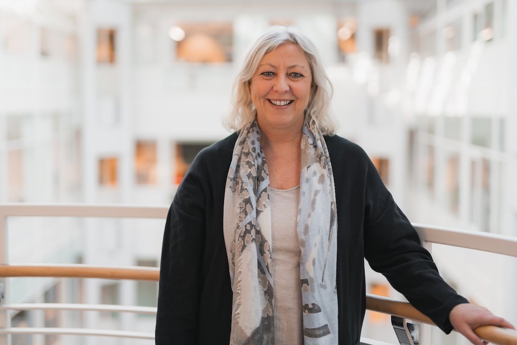 En glad eldre kvinne med blondt, skulderlangt hår står på en gangbro i en moderne bygning.