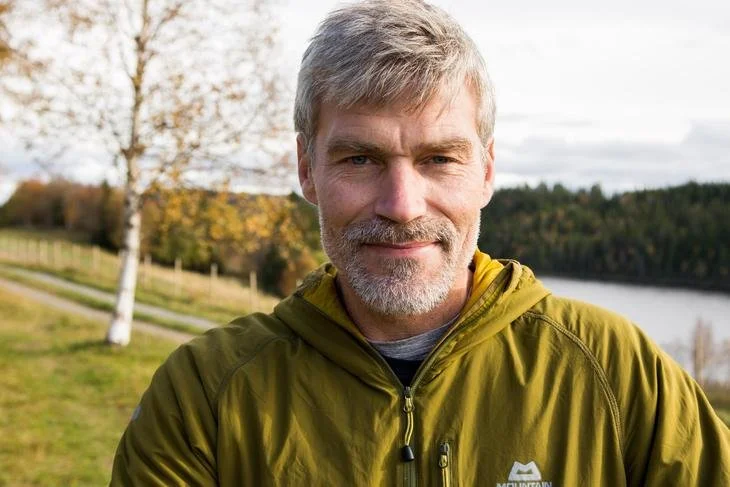 En gammel mann med grått hår og skjegg smiler, iført en gul jakke, ute i naturen med trær og en innsjø i bakgrunnen.