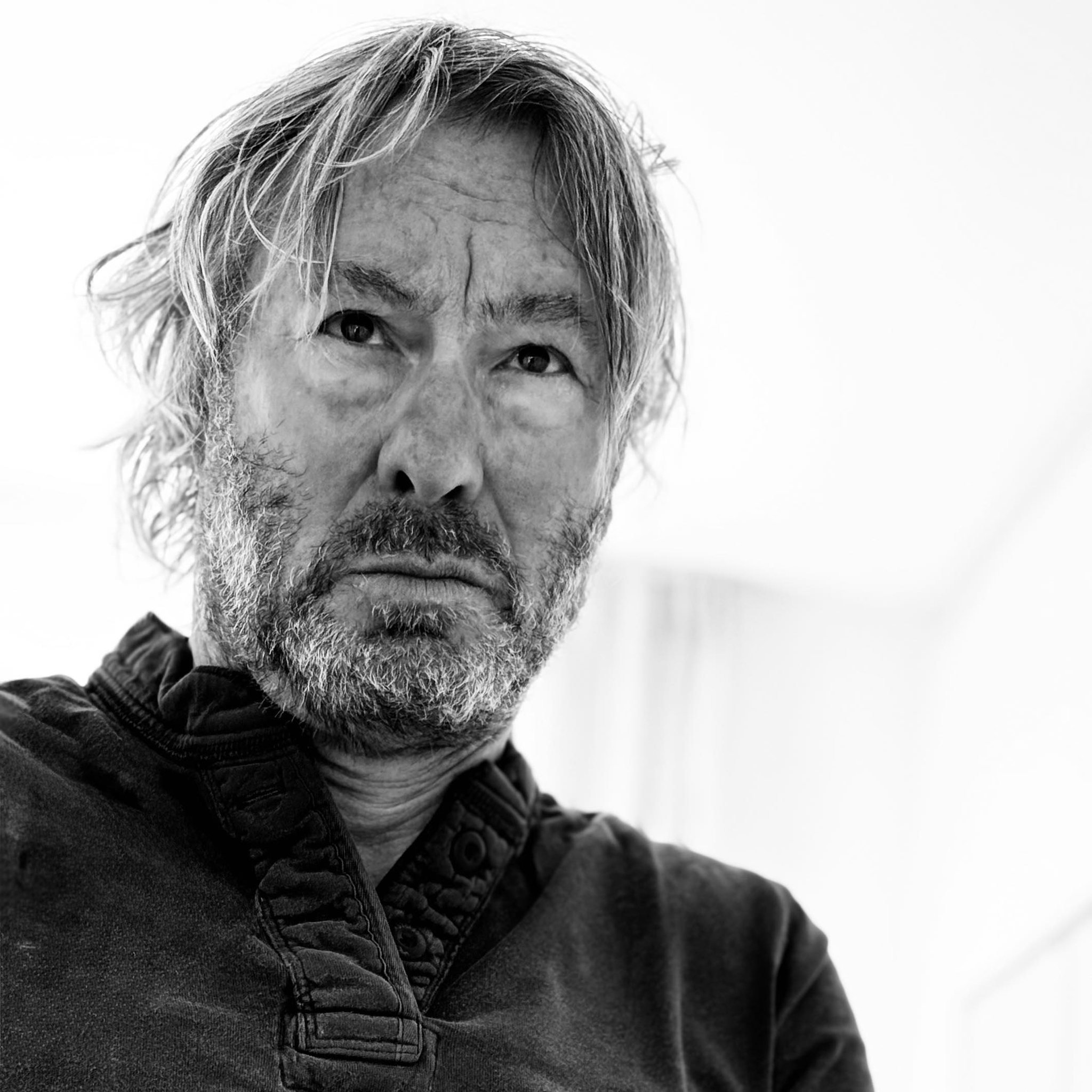Portrett av en eldre mann med grått hår og skjegg, iført en svart jakke, fotografert i svart-hvitt.