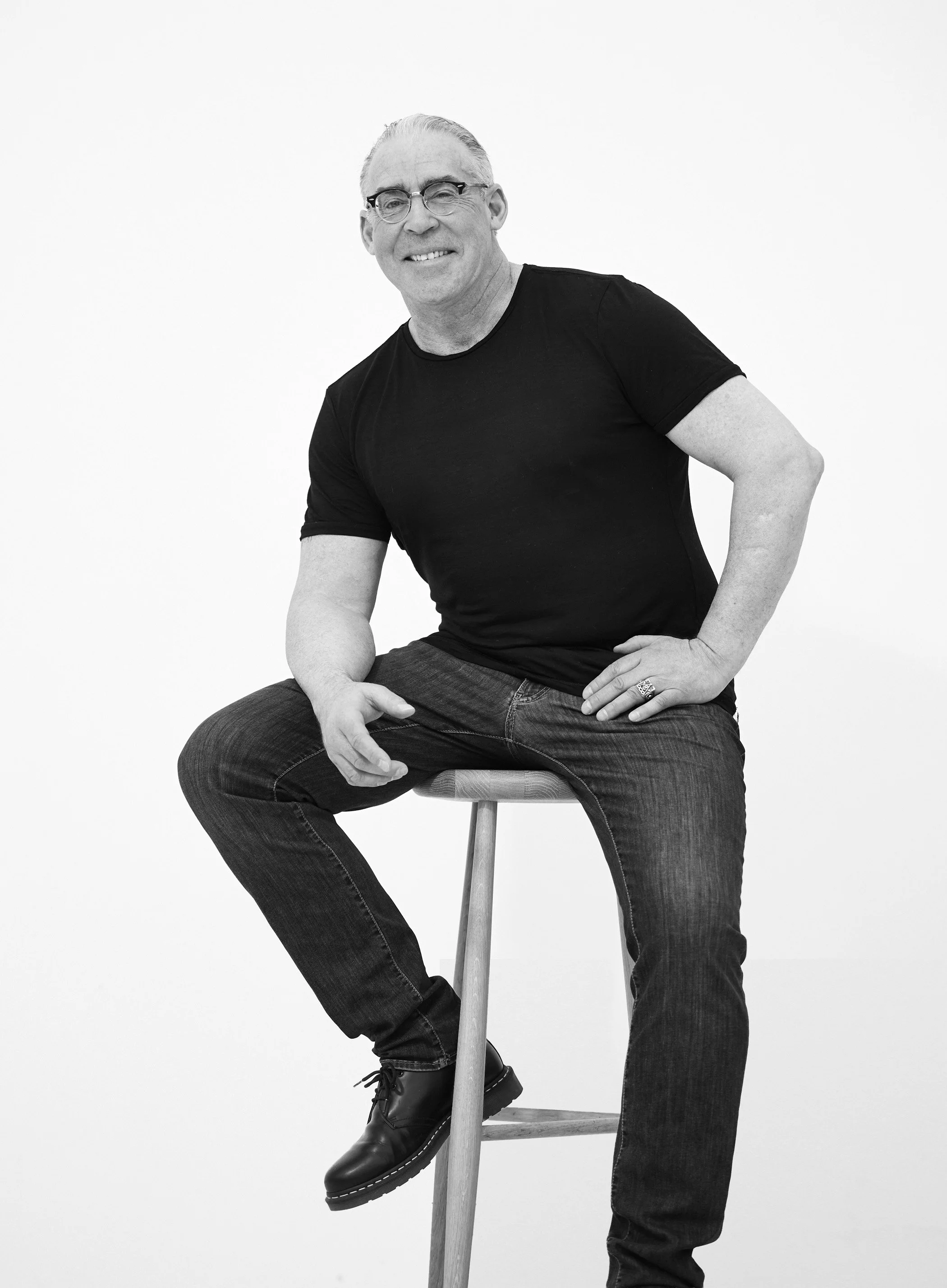 En mann med briller og lyst hår sitte på en stol, smiler mot kameraet, iført svart t-skjorte og jeans, med en stor hvit bakgrunn.