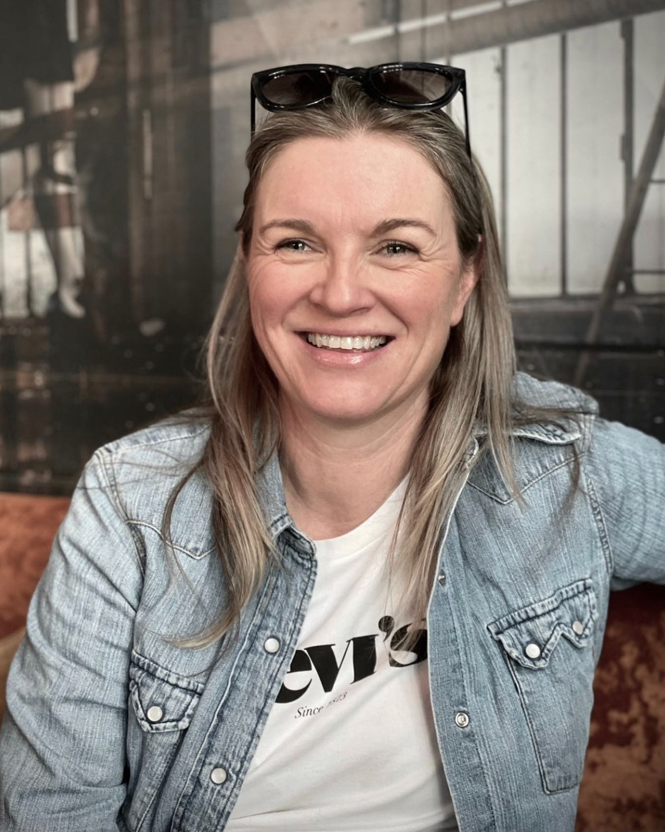 En smilende kvinne med langt, blond hår, iført en lys denimjakke og en hvit T-skjorte, sitter innendørs, med et bilde av en barneroms- eller stuebakgrunn.