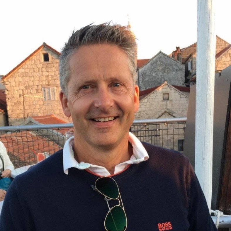 En smilende mann med kort grått hår, iført en hvit skjorte og en mørk genser, står utendørs med hus i bakgrunnen.