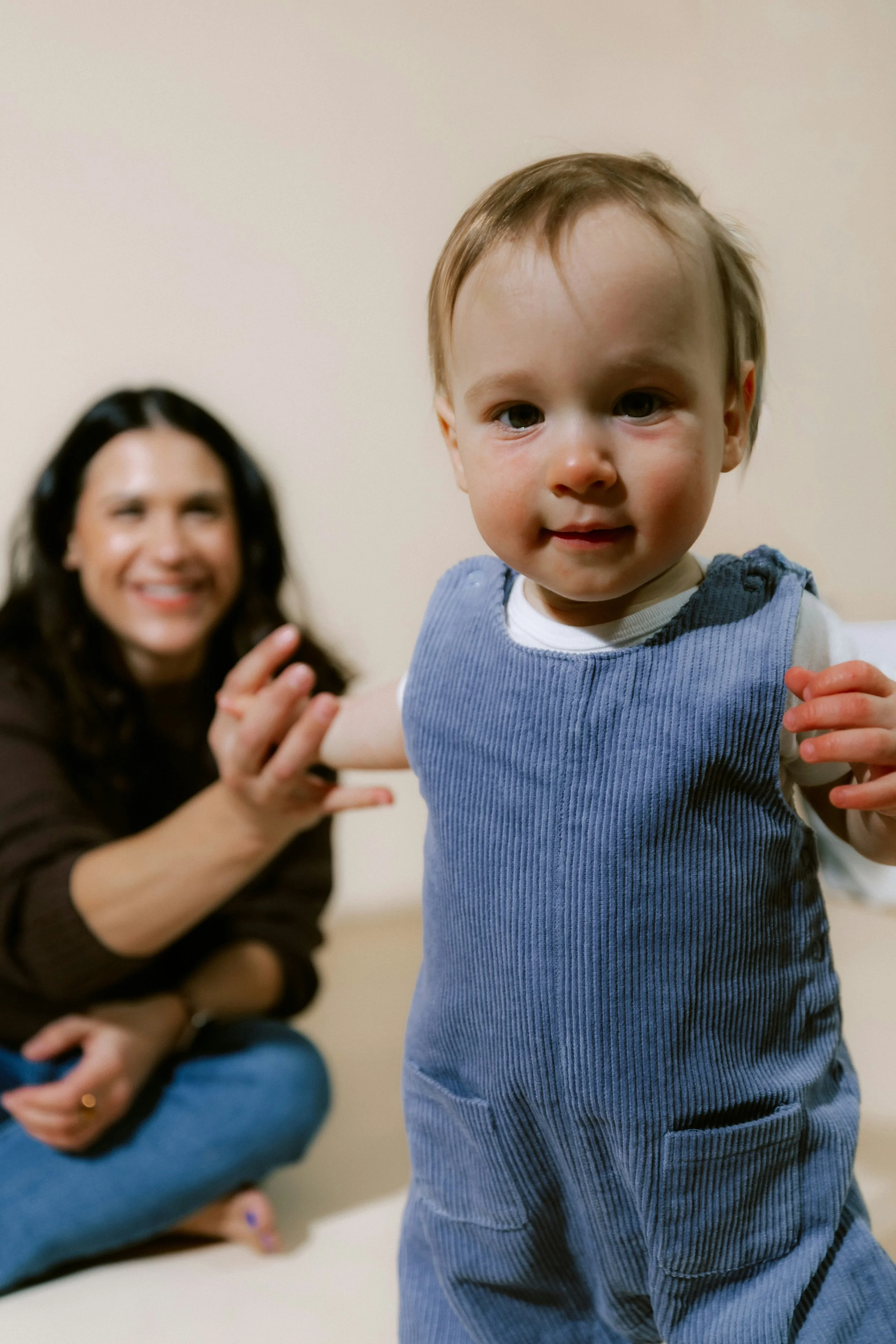 A young boy in blue overalls is in the foreground, looking directly at the camera with a slight smile. Behind him, a woman with long dark hair, sitting cross-legged, is smiling and holding the boy's arm.