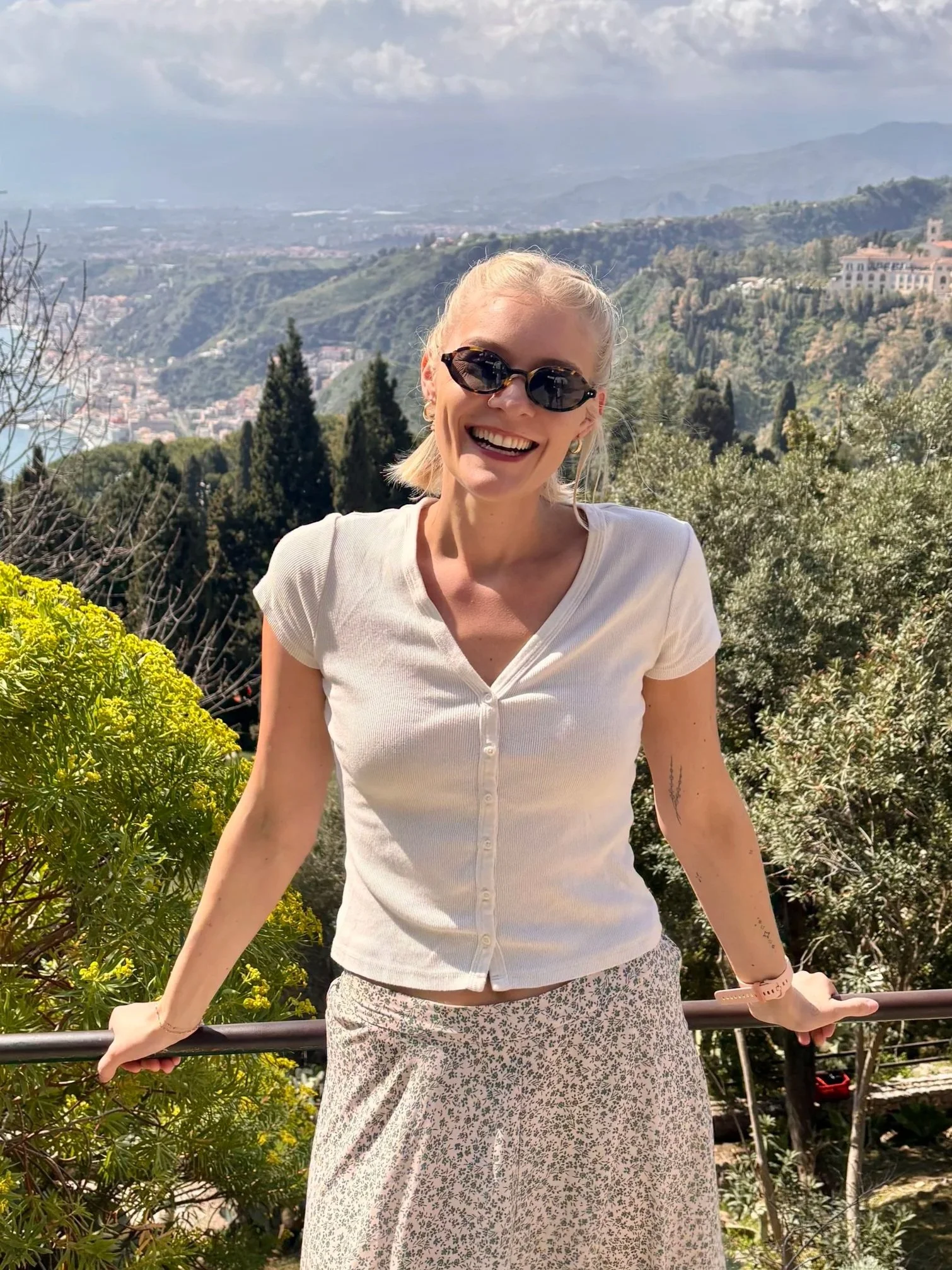 A woman with blonde hair, wearing sunglasses, a white button-up shirt, and a floral skirt, standing outdoors with a scenic mountainous landscape in the background.