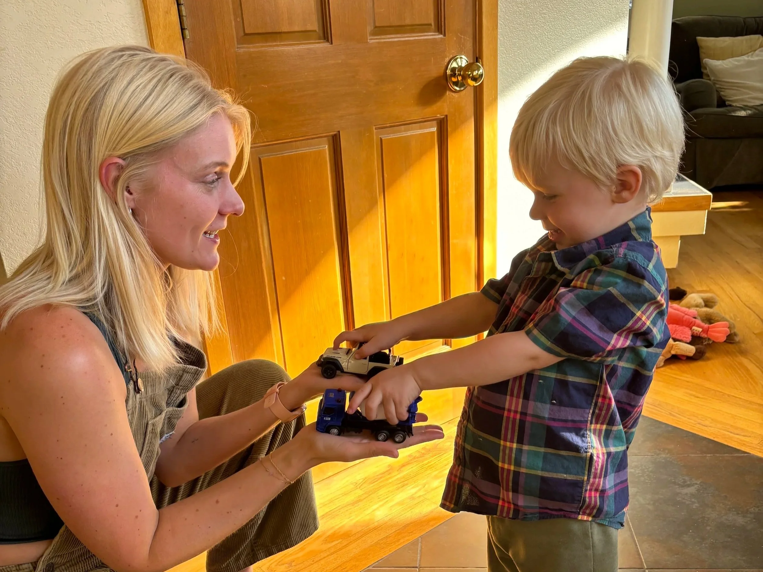 A young boy with blonde hair, wearing a plaid shirt, and a woman with blonde hair, smiling, sharing and playing with toy cars inside a home, near a wooden door and hardwood floor.