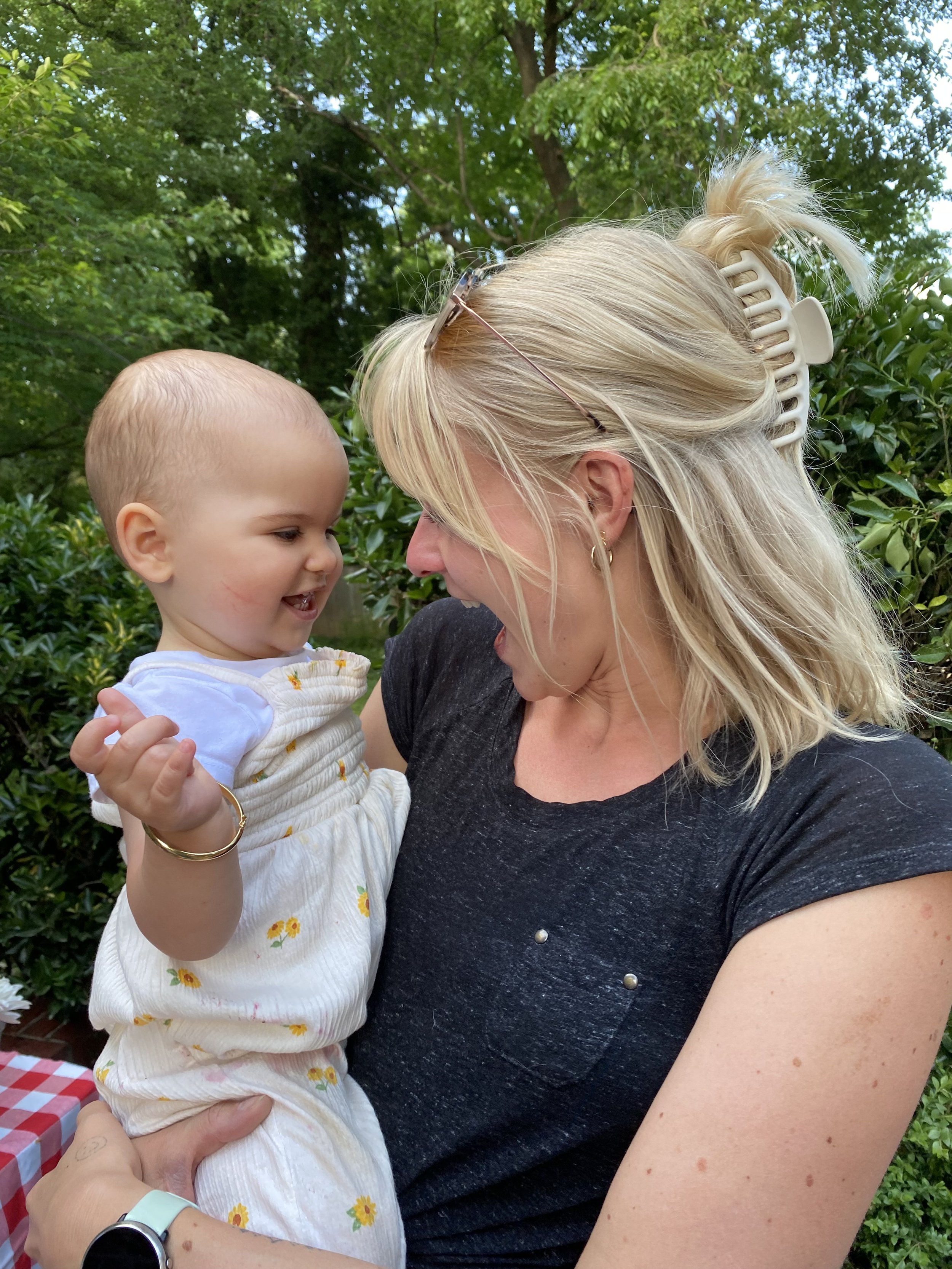 A woman with blonde hair holding a laughing young child outdoors surrounded by green trees and bushes.