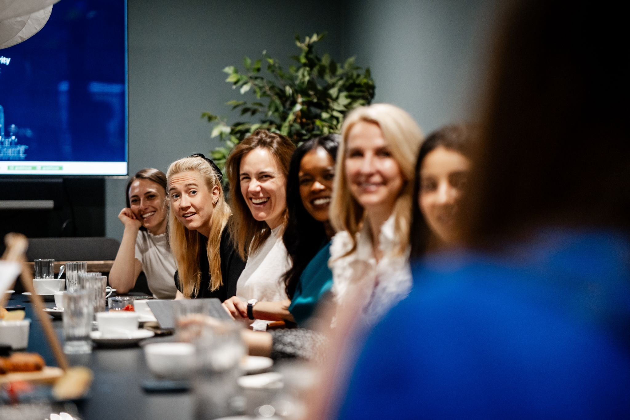 A group of women sitting at a table during a meeting or conference, smiling, with food and drinks on the table, and a large screen visible on the wall in the background.